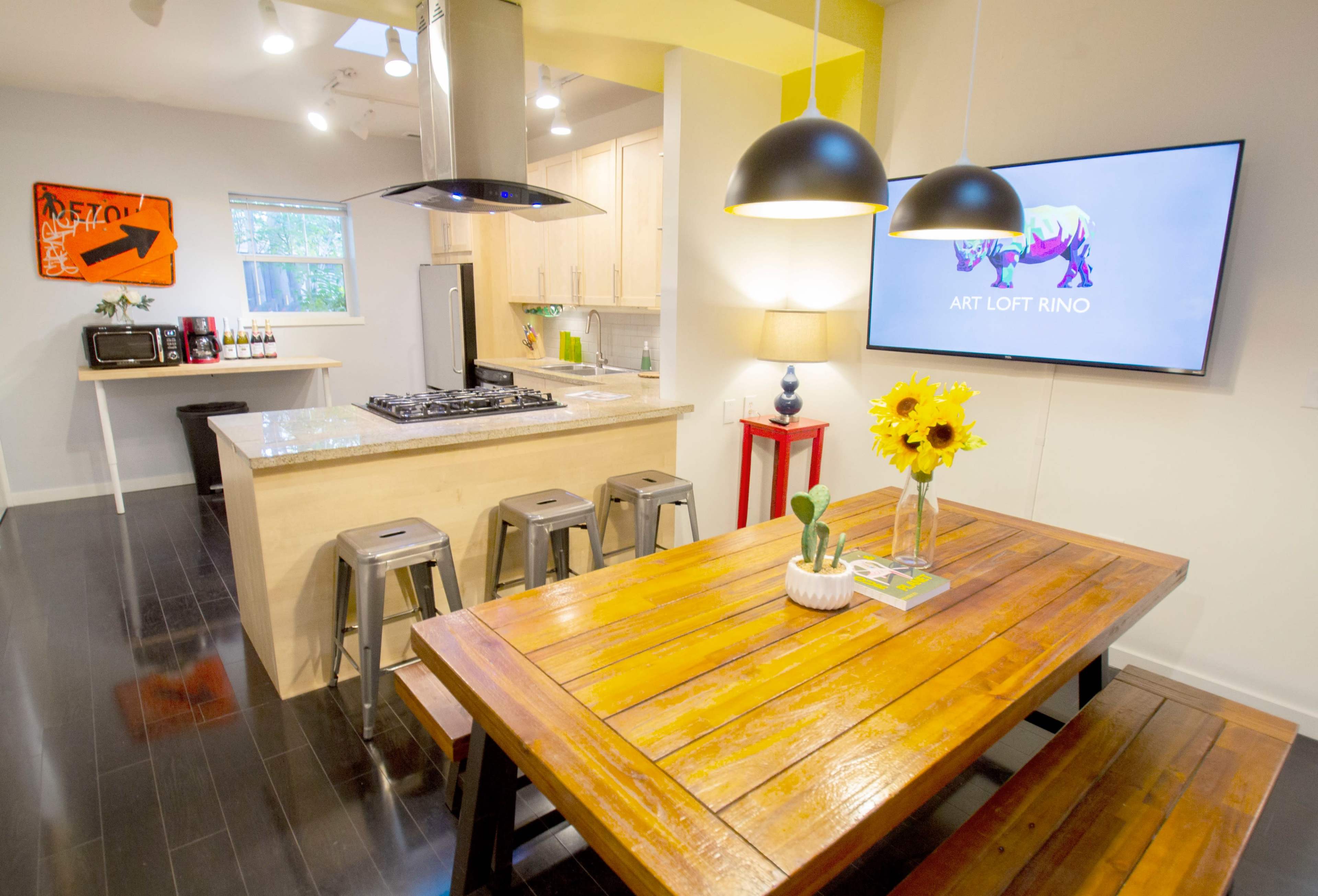 The image shows a modern kitchen and dining area with a wooden table, bar stools, and a large television displaying the words "ART LOFT RINO."
