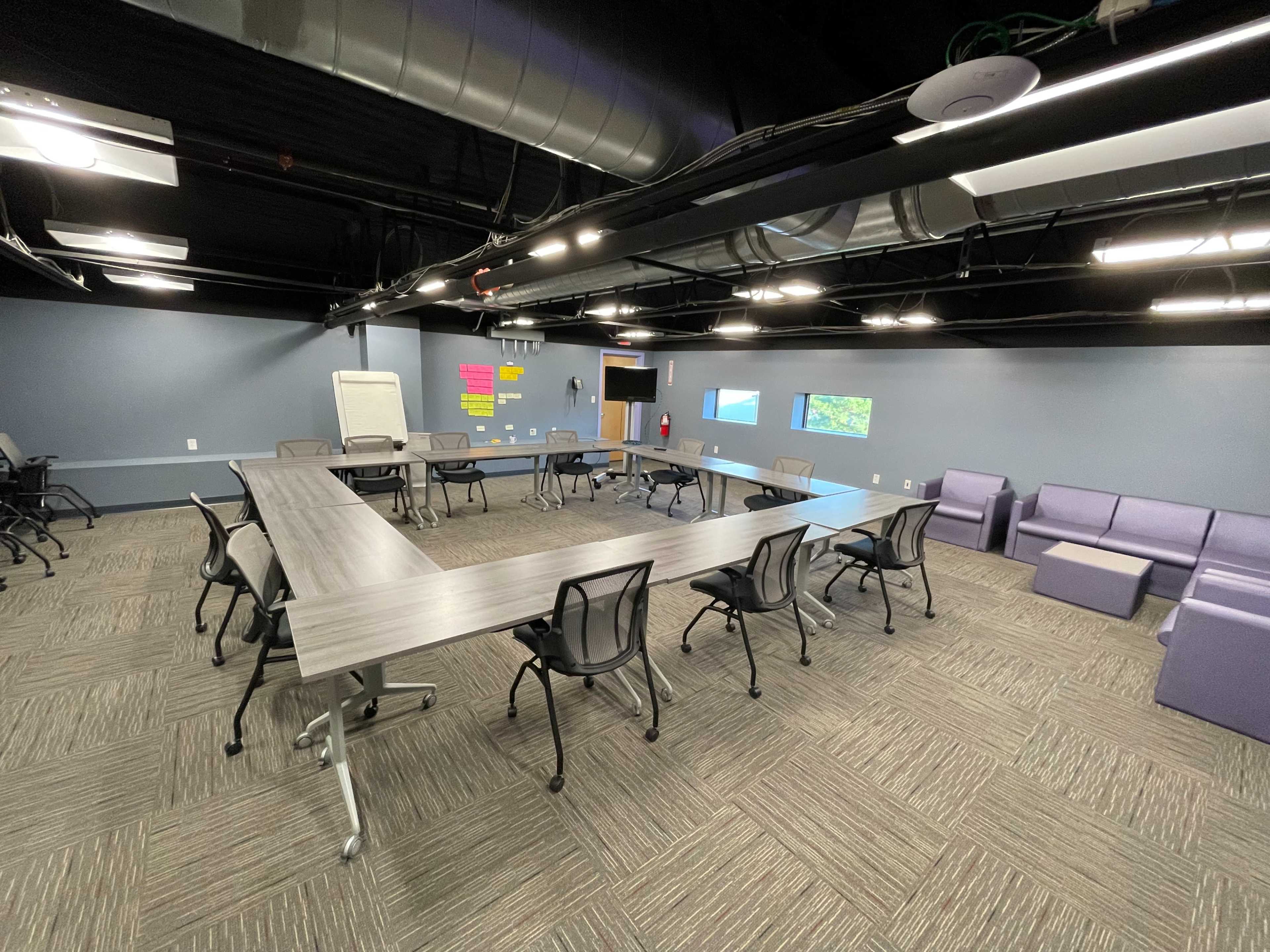 A meeting room features a large rectangular table surrounded by rolling chairs, with a purple couch and a whiteboard in the background.