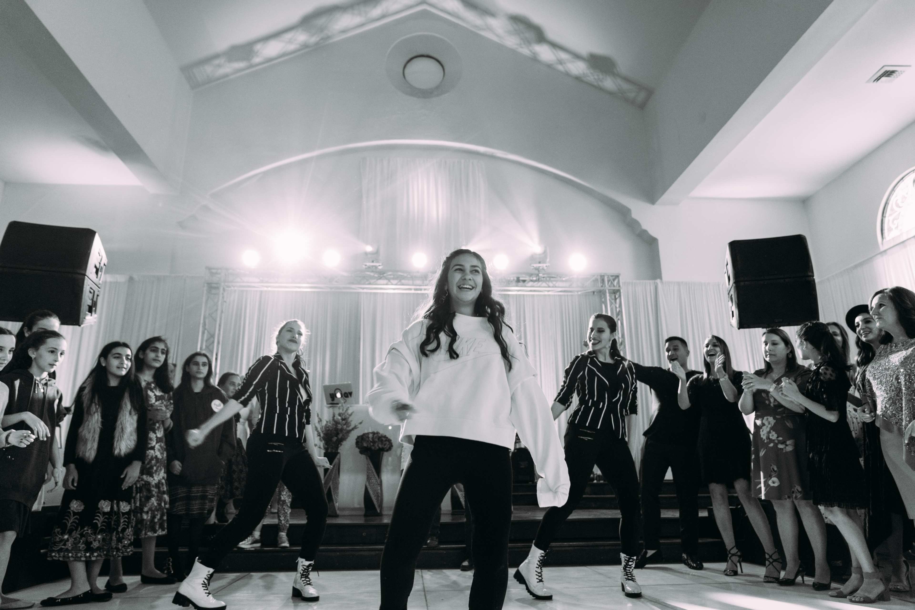 A group of dancers performs on stage while a crowd observes and applauds in a well-lit event space.