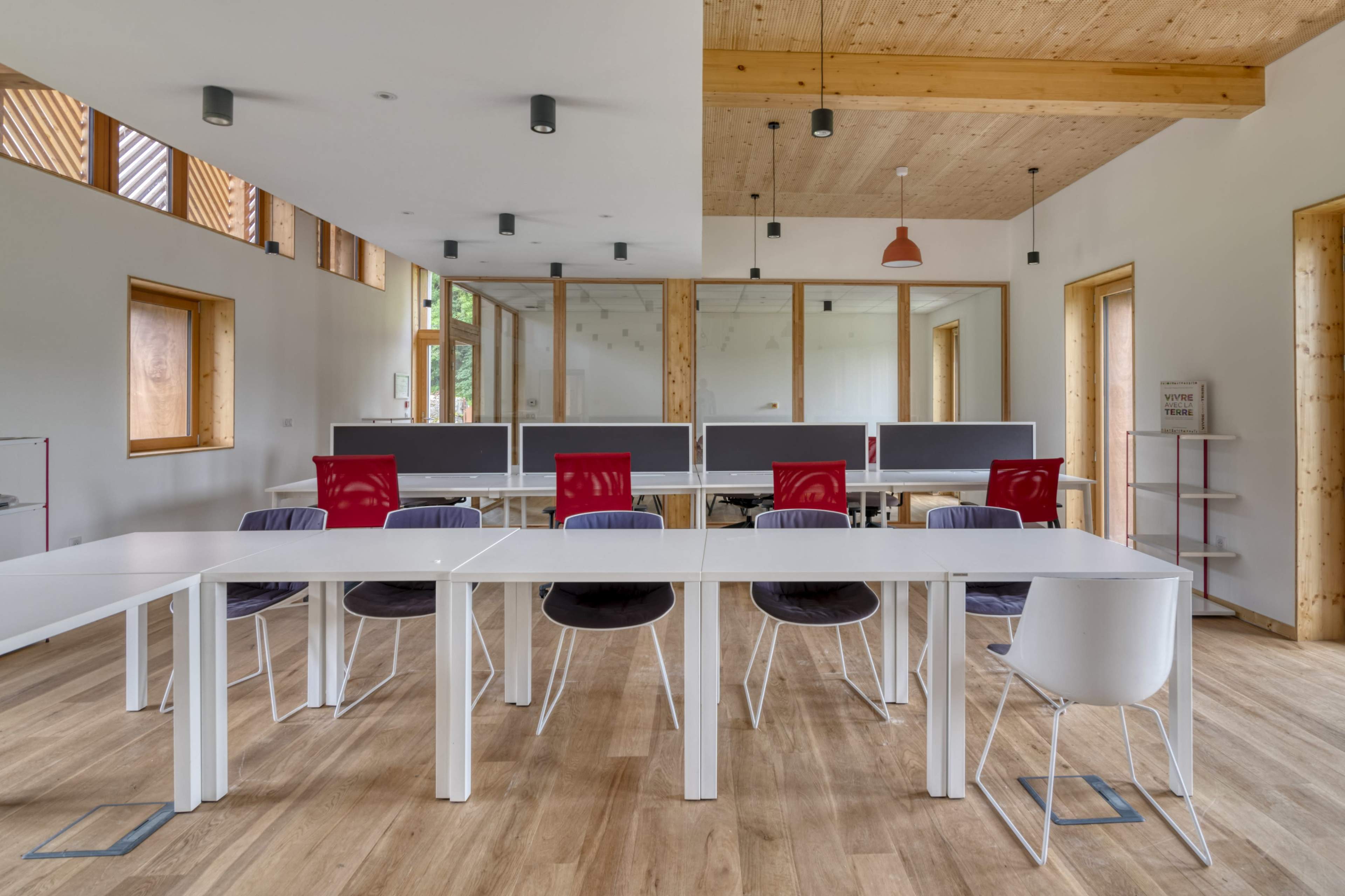 A modern office space features several rows of white desks with computers, surrounded by wooden walls and large windows, creating an open and airy atmosphere.