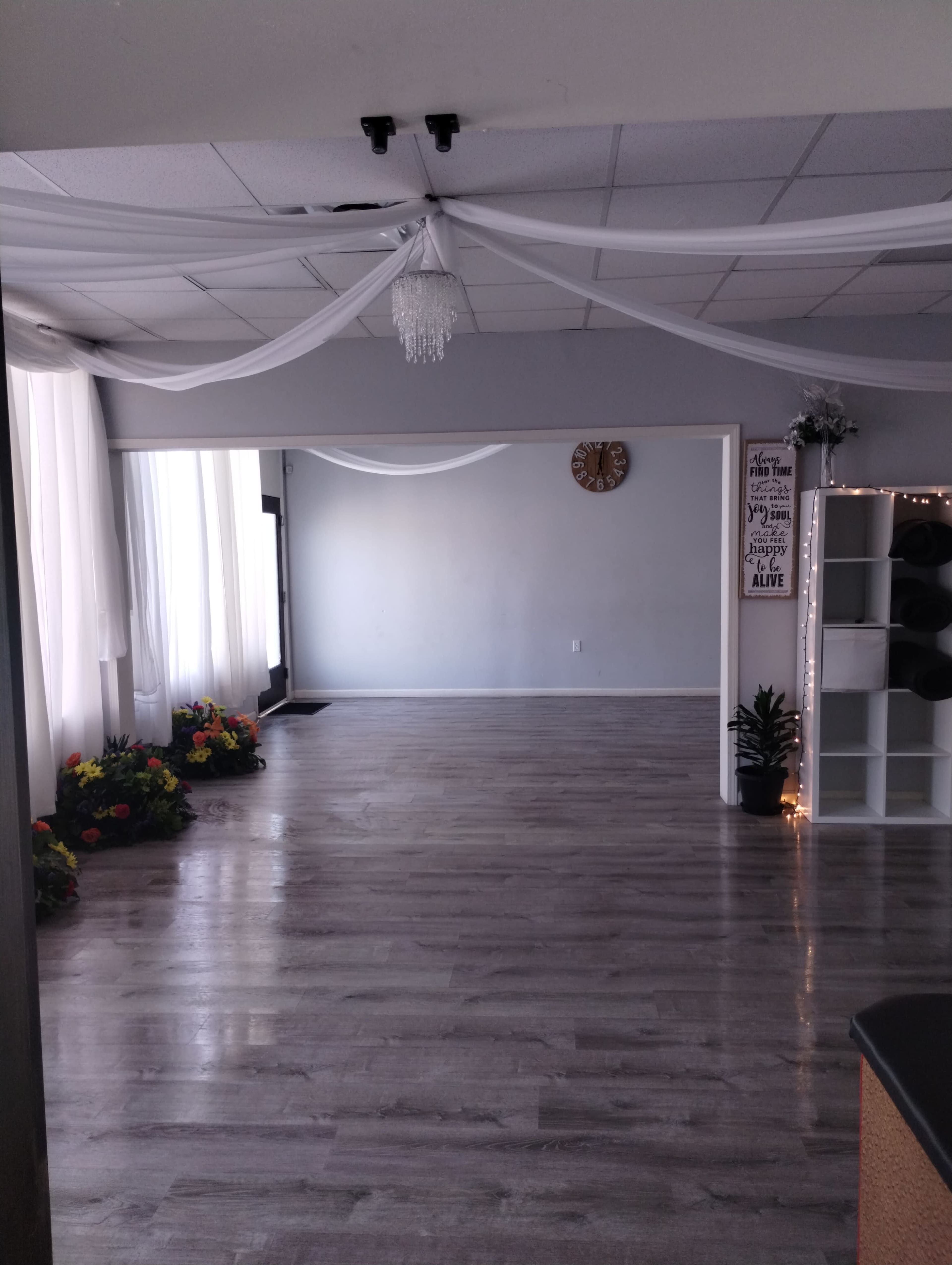A spacious room with large windows, light-colored wooden flooring, and decorations including a clock and potted plants.
