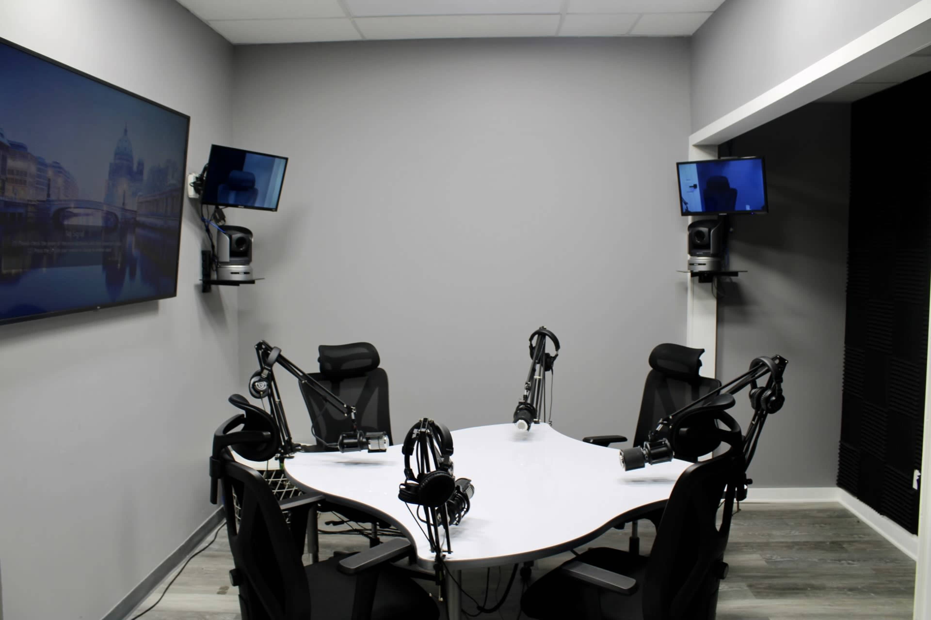 The image shows a modern podcast recording studio with a white table, multiple microphones, and two monitors mounted on the walls.
