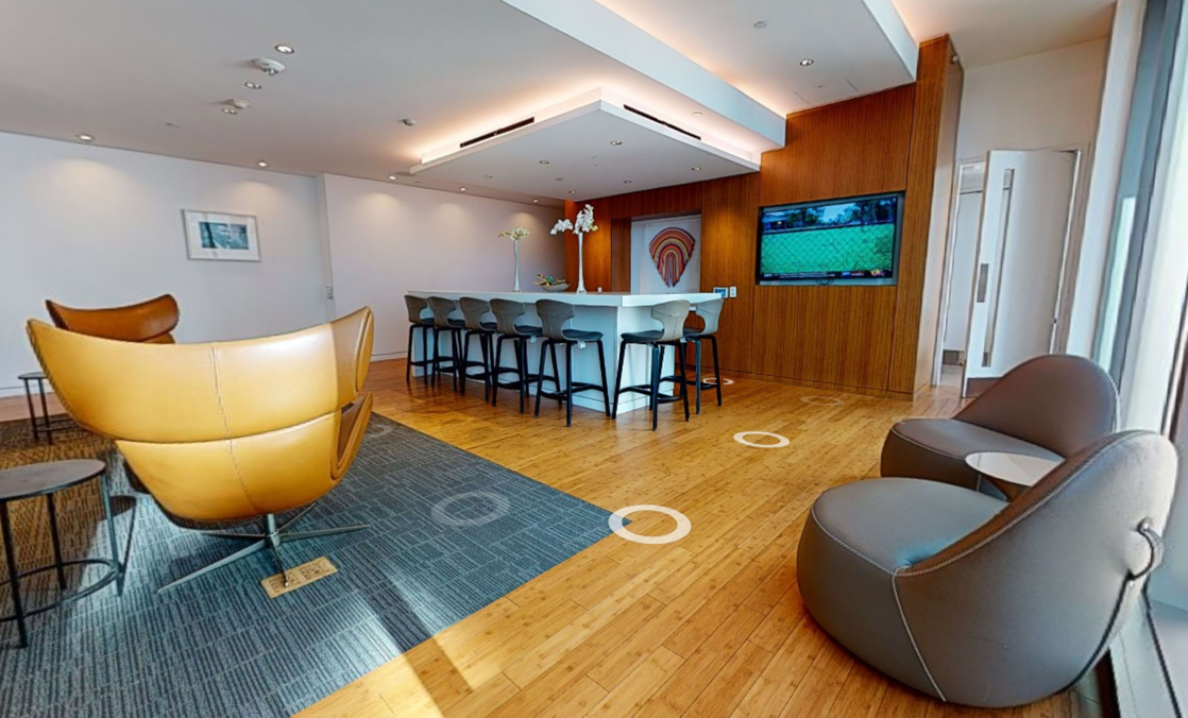 A modern lounge area with a bar counter, several seating options, and a large television displaying a sports broadcast.