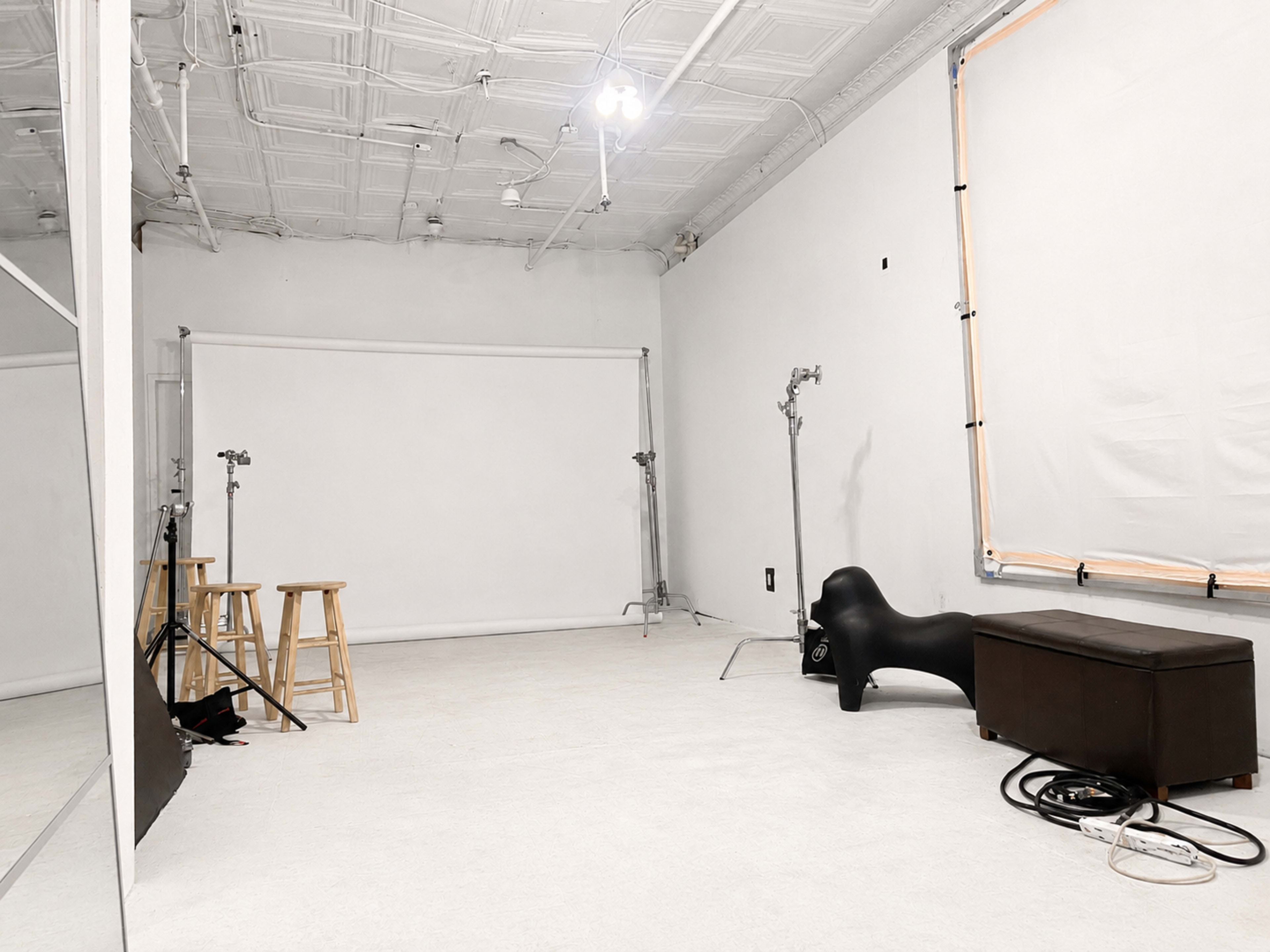 The image shows a spacious studio with white walls, a light-colored floor, and equipment including two wooden stools, a black bench, and lighting stands.