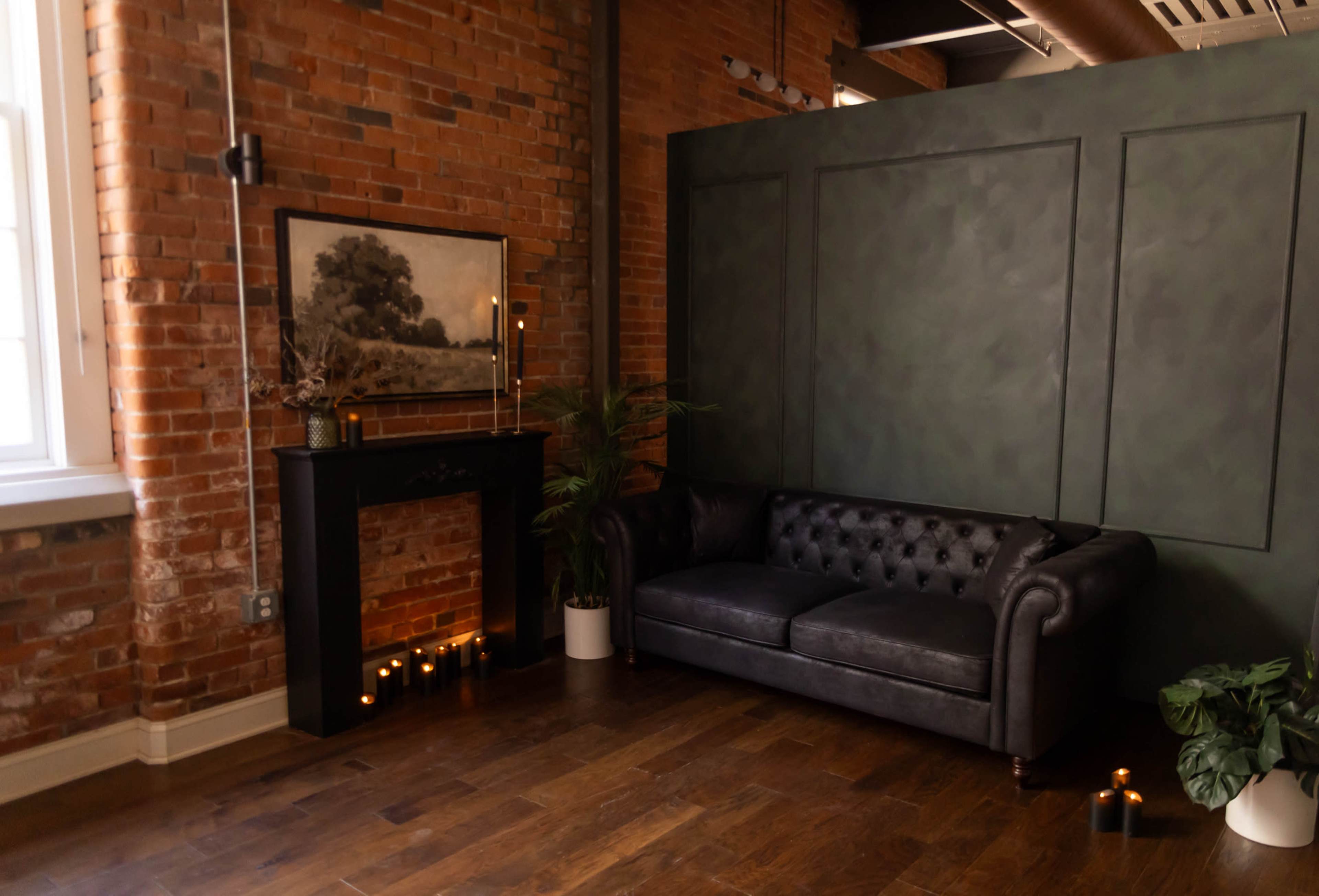 The image shows a cozy interior with a dark leather couch, a brick wall, a green accent wall, and candles arranged on the floor near a fireplace.