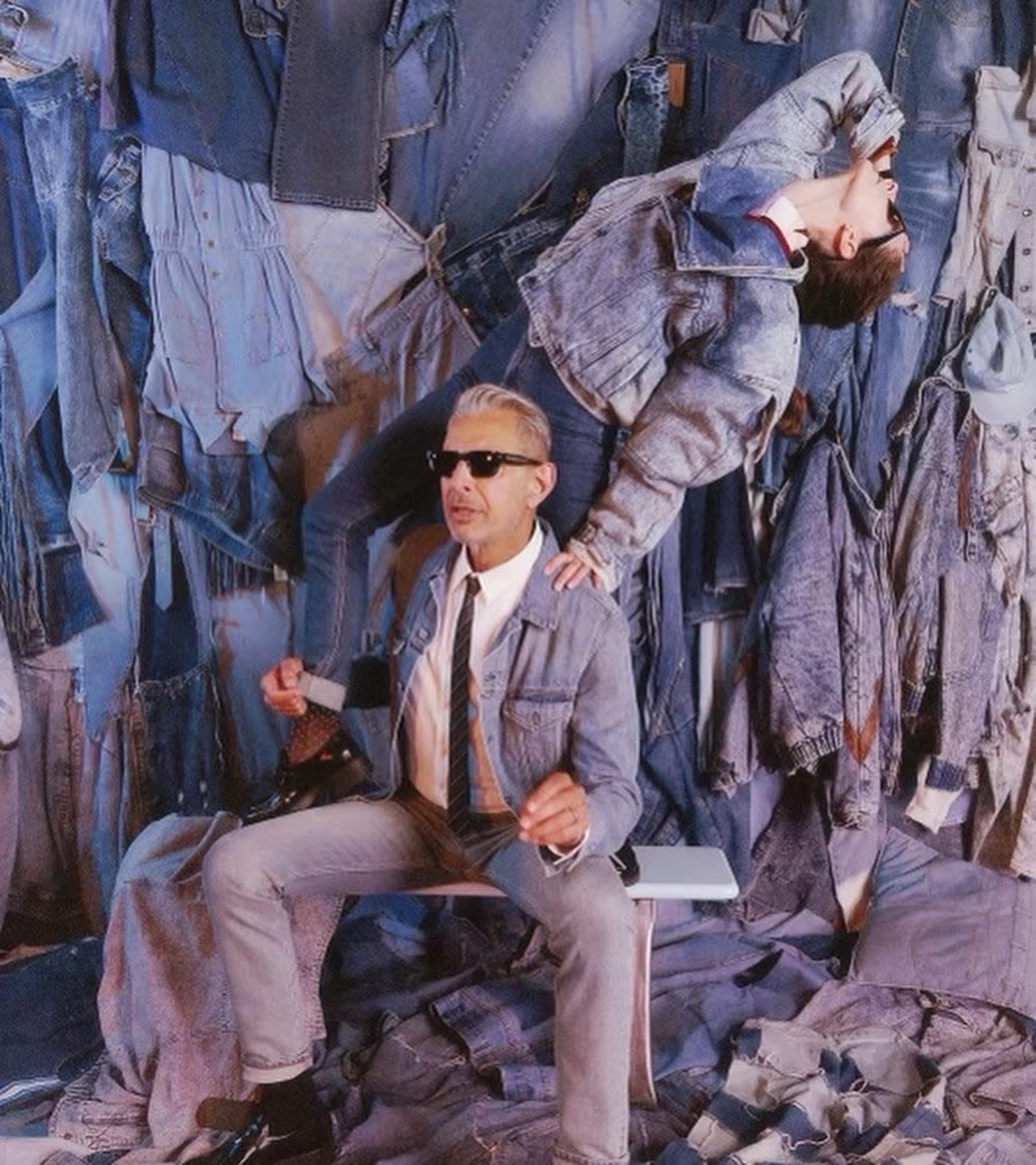 A man in sunglasses sits on a chair amidst a backdrop of blue denim, while a second person is dramatically posed upside-down behind him.