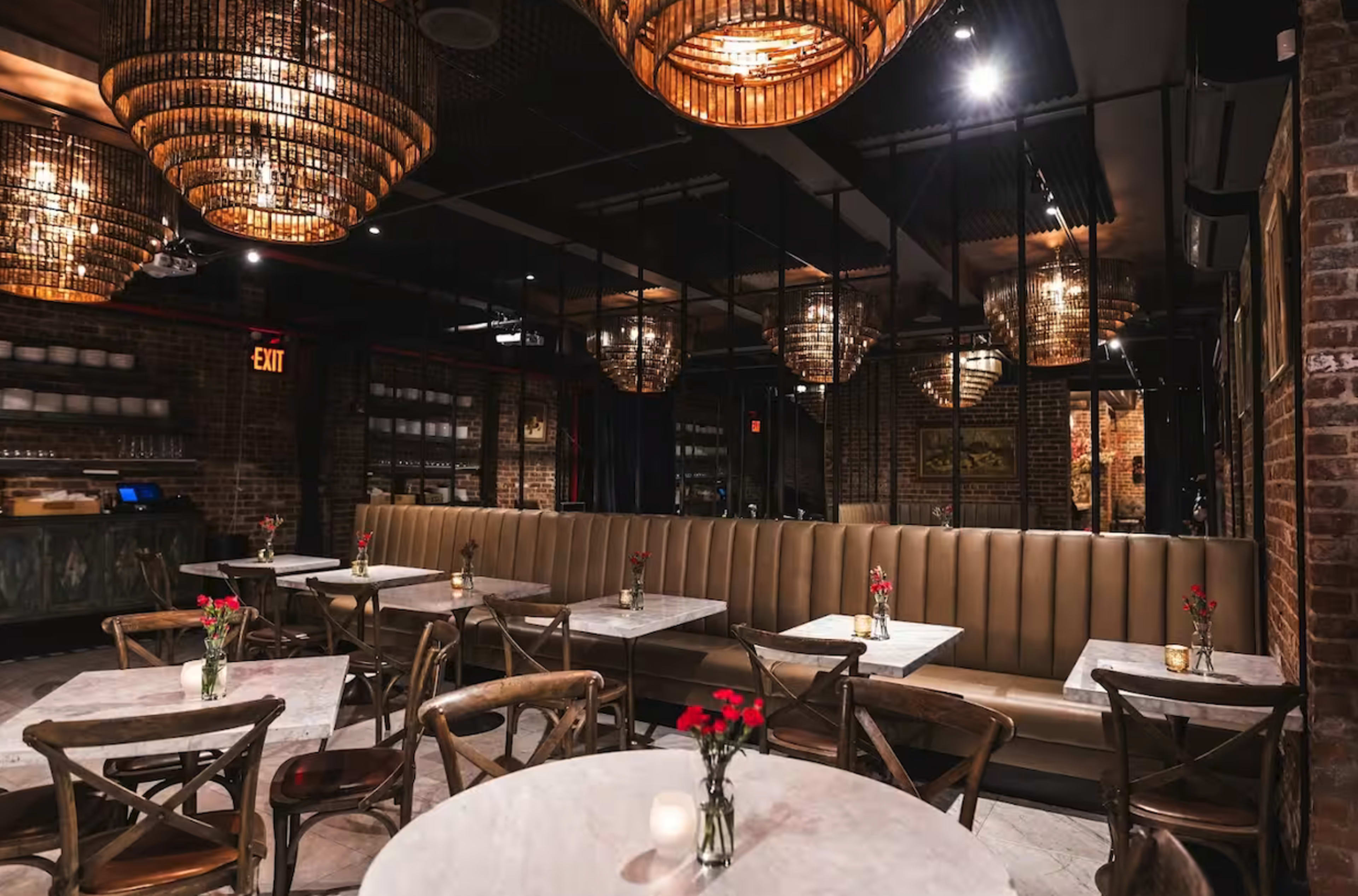 The image shows an interior view of a restaurant featuring beige banquettes, round marble tables, and decorative pendant lights hanging from the ceiling.