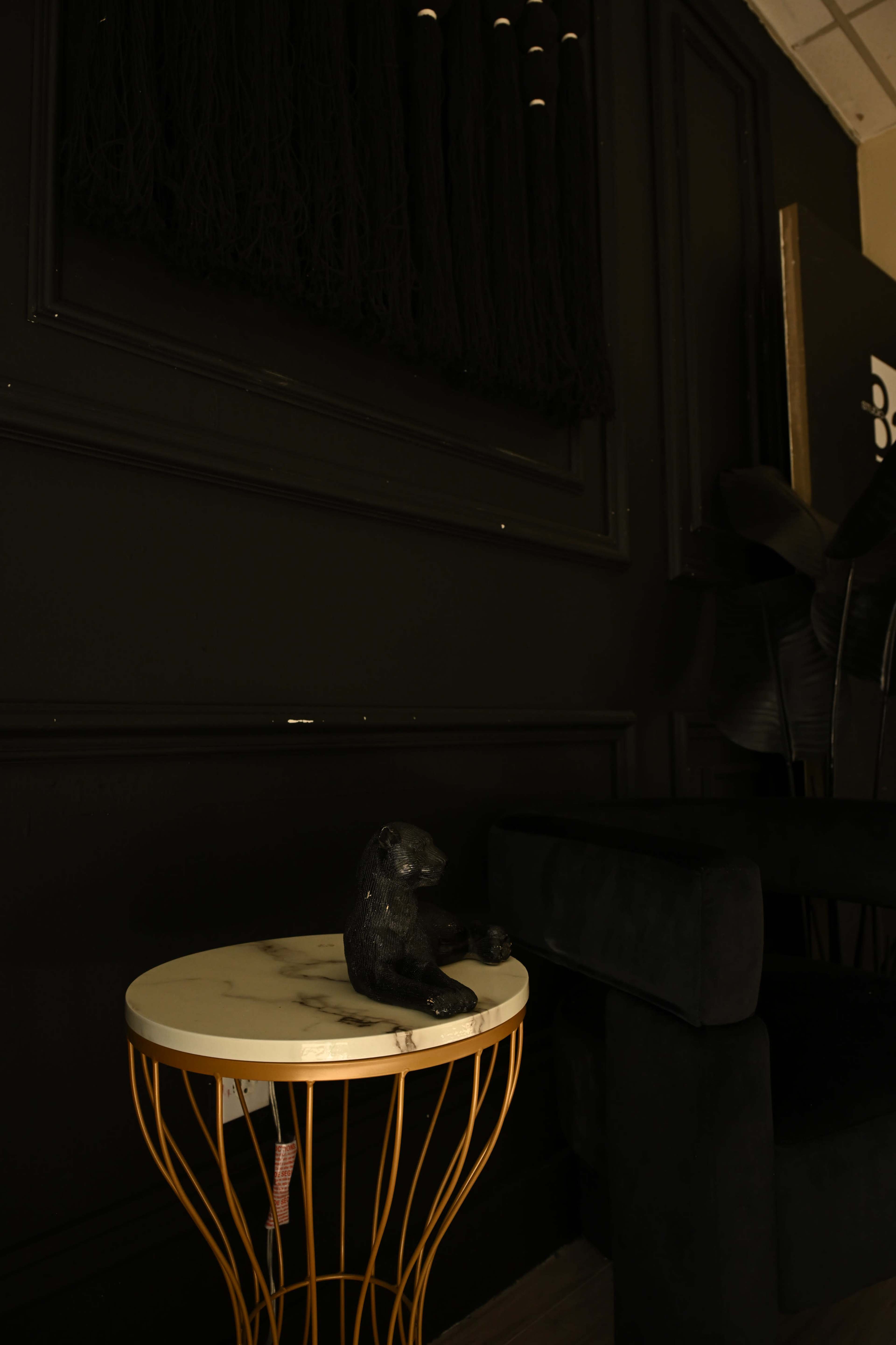 A black decorative figurine of an animal sits on a marble-topped table with a gold wire base, against a dark wall and a plush black sofa.