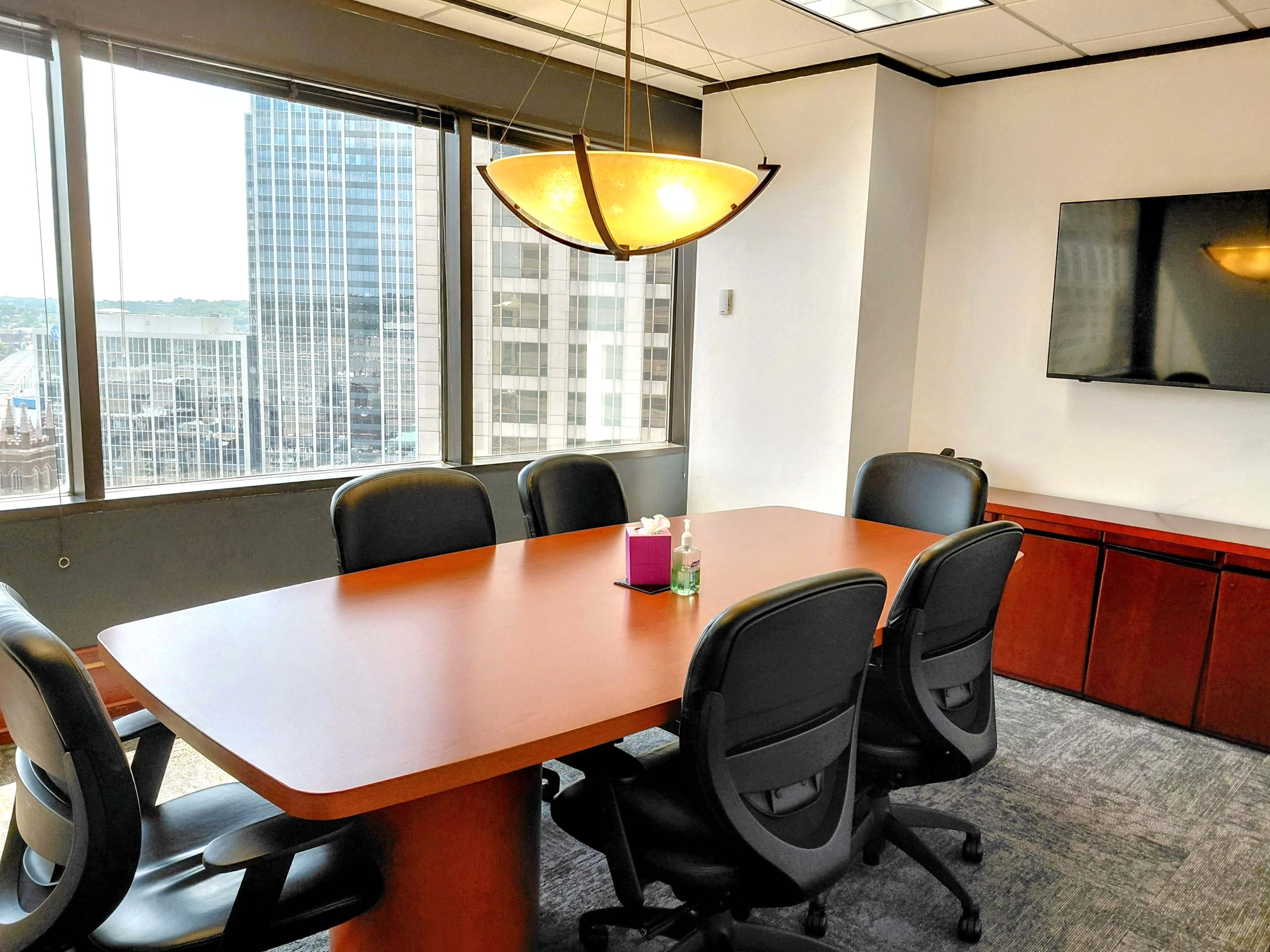 The image shows a modern conference room with a large wooden table, black chairs, and a window offering a view of city buildings.