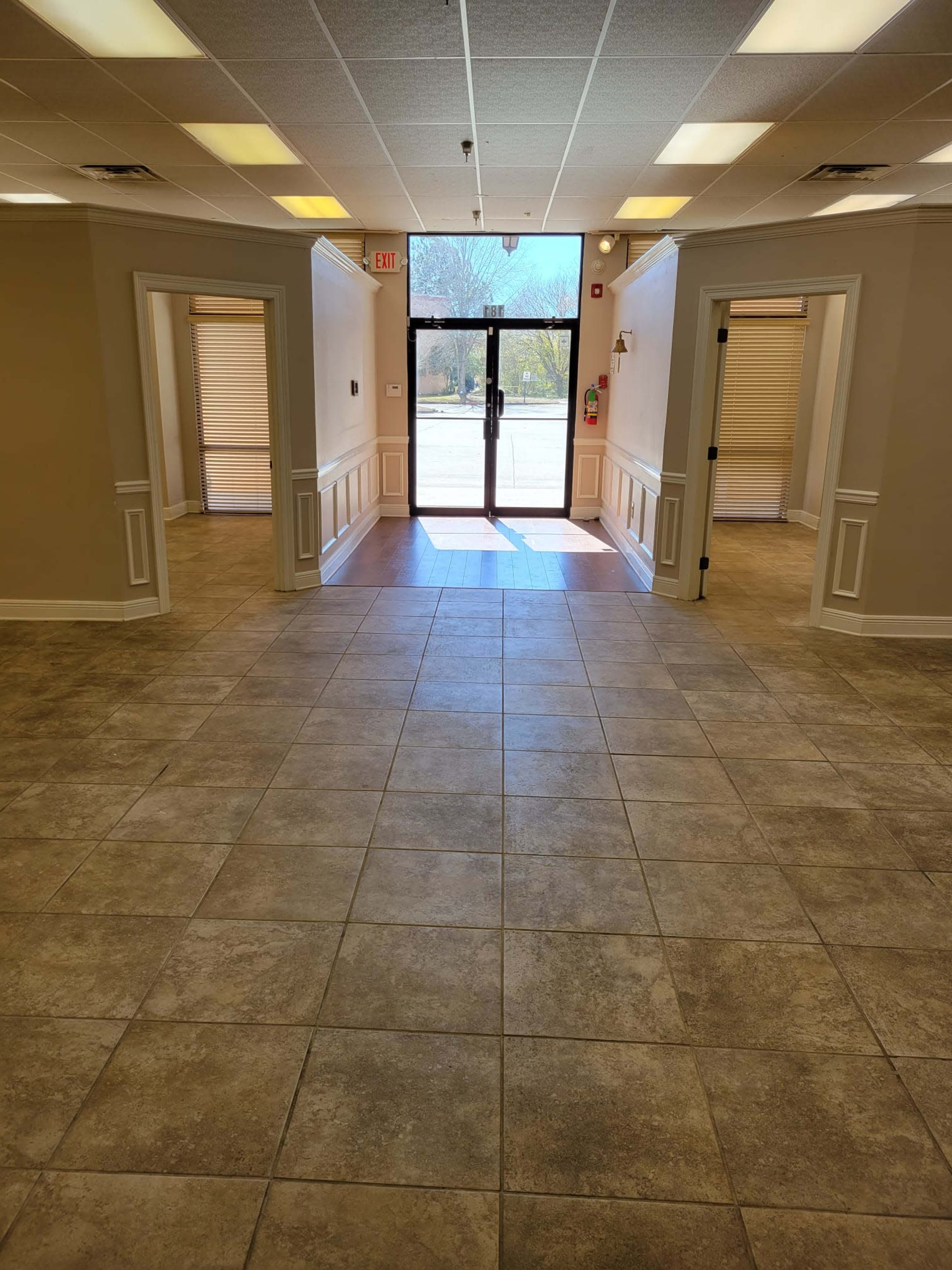A spacious corridor with tile flooring leading to a glass entryway, flanked by doorways on either side.