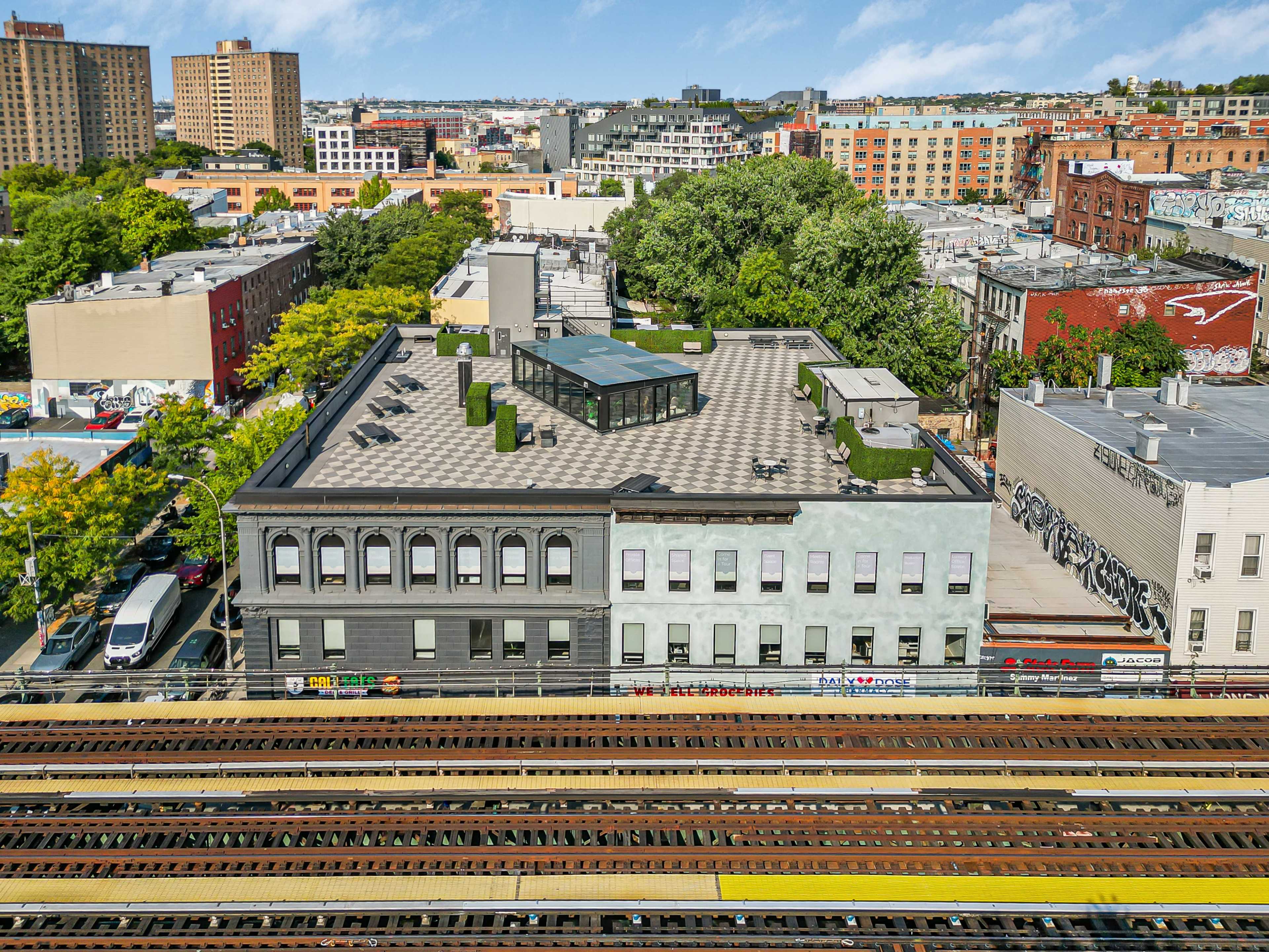 The image shows a rooftop view of a building adjacent to elevated train tracks, surrounded by urban scenery that includes other buildings and trees.