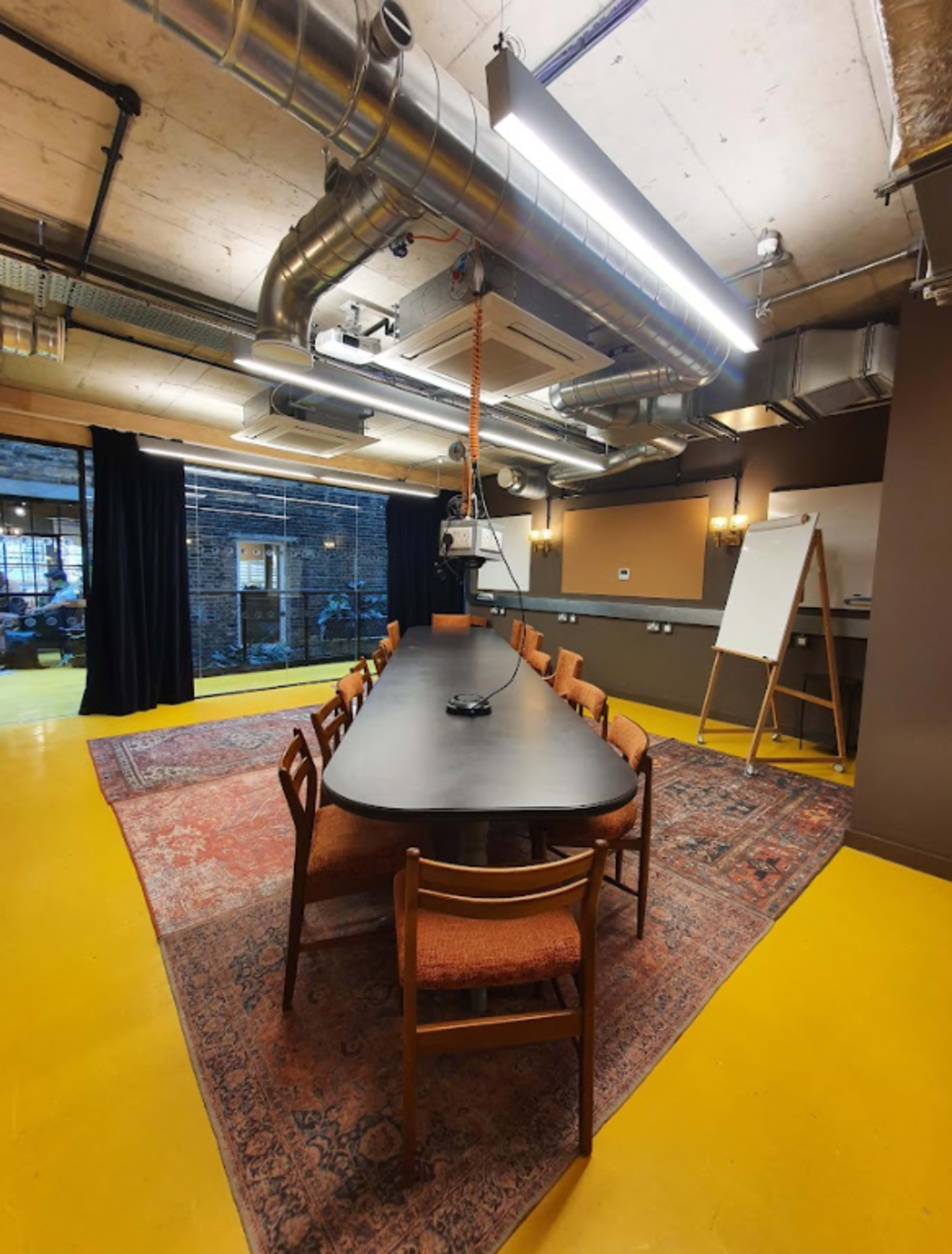 A long rectangular table with wooden chairs is set in a meeting room featuring industrial-style decor and a yellow floor.