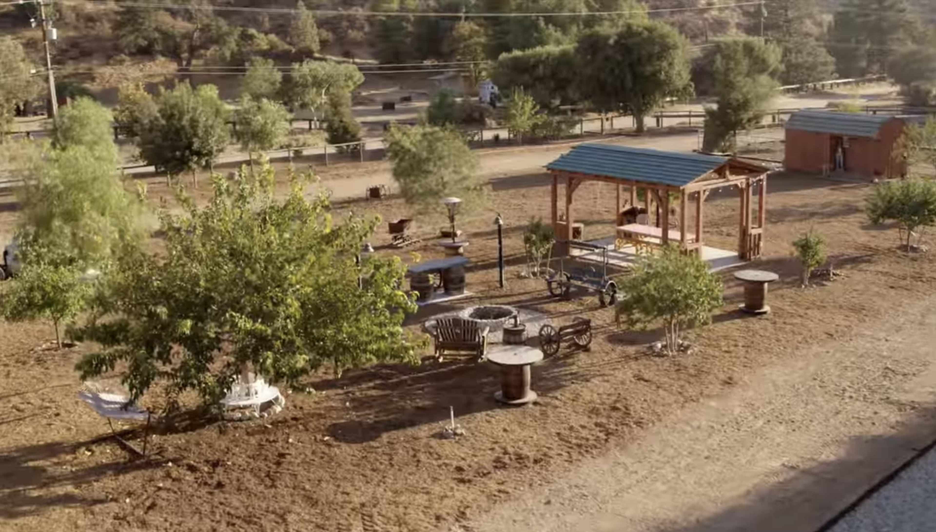 The image shows an outdoor area with wooden structures, including a covered gazebo, surrounded by trees and garden furniture.