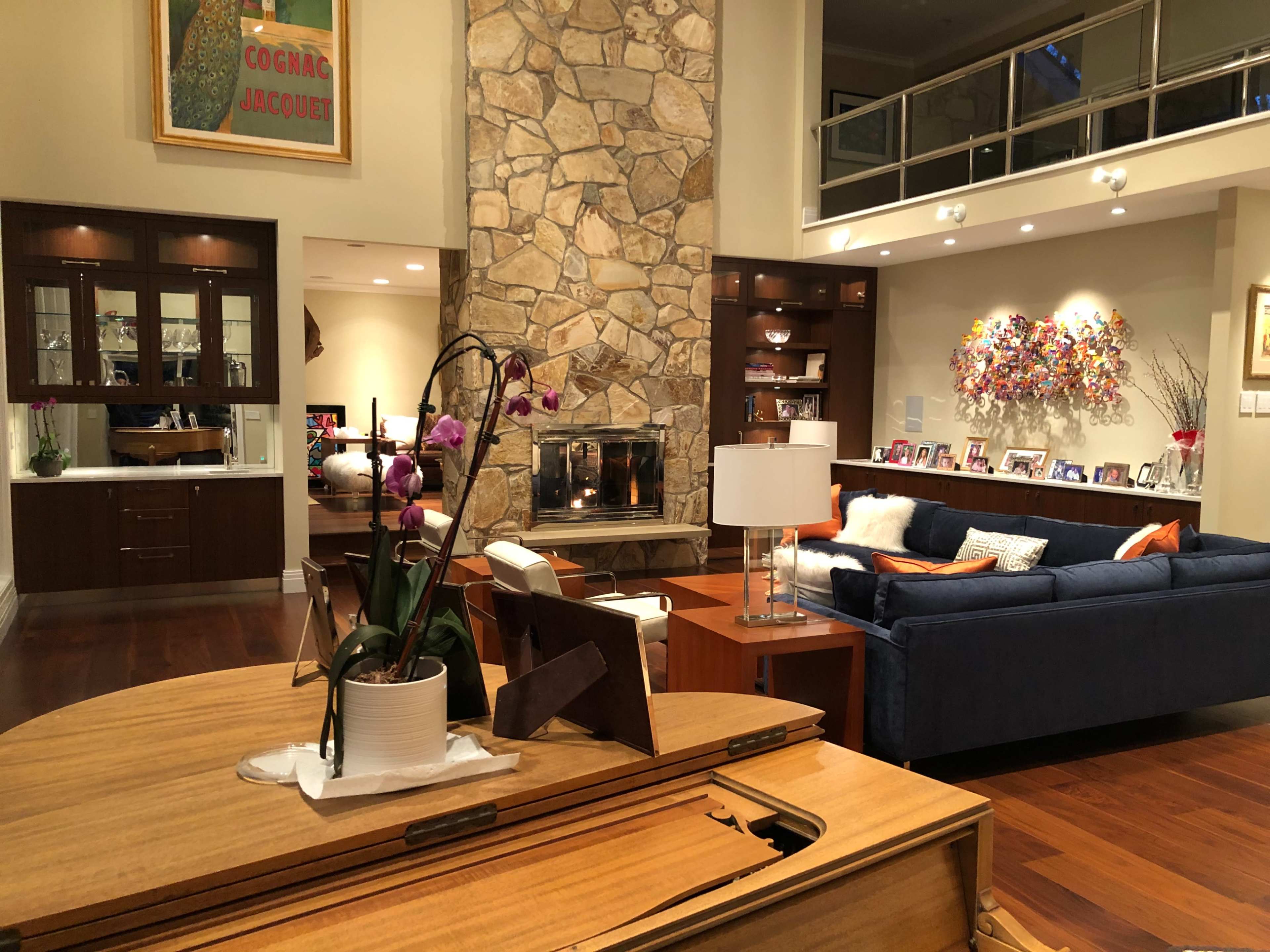 The image shows a spacious living room featuring a stone fireplace, a blue sofa, a wooden piano in the foreground, and decorative elements including a floral arrangement and framed photos on the walls.