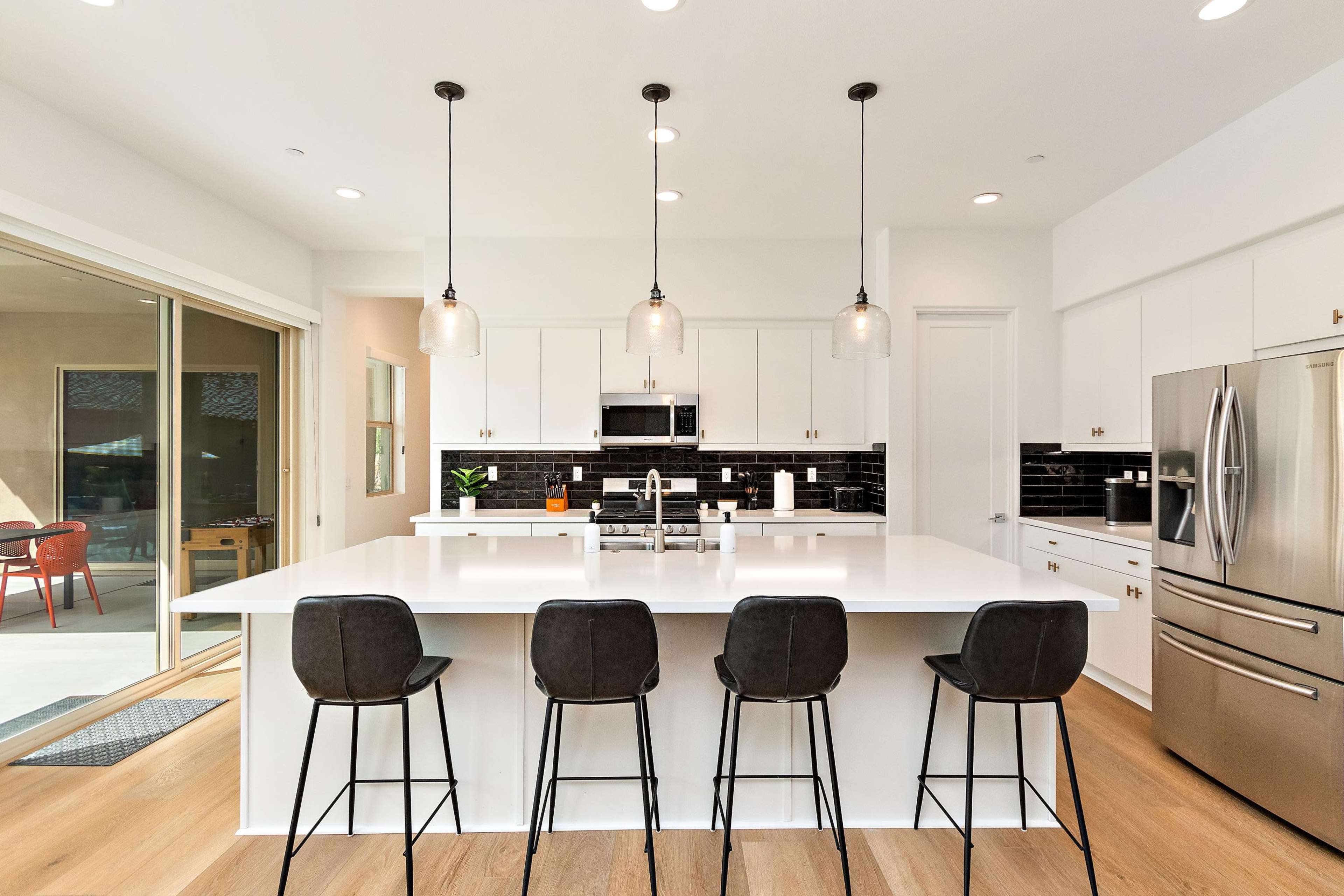 A modern kitchen featuring a large island with four high stools, white cabinetry, stainless steel appliances, and pendant lighting.
