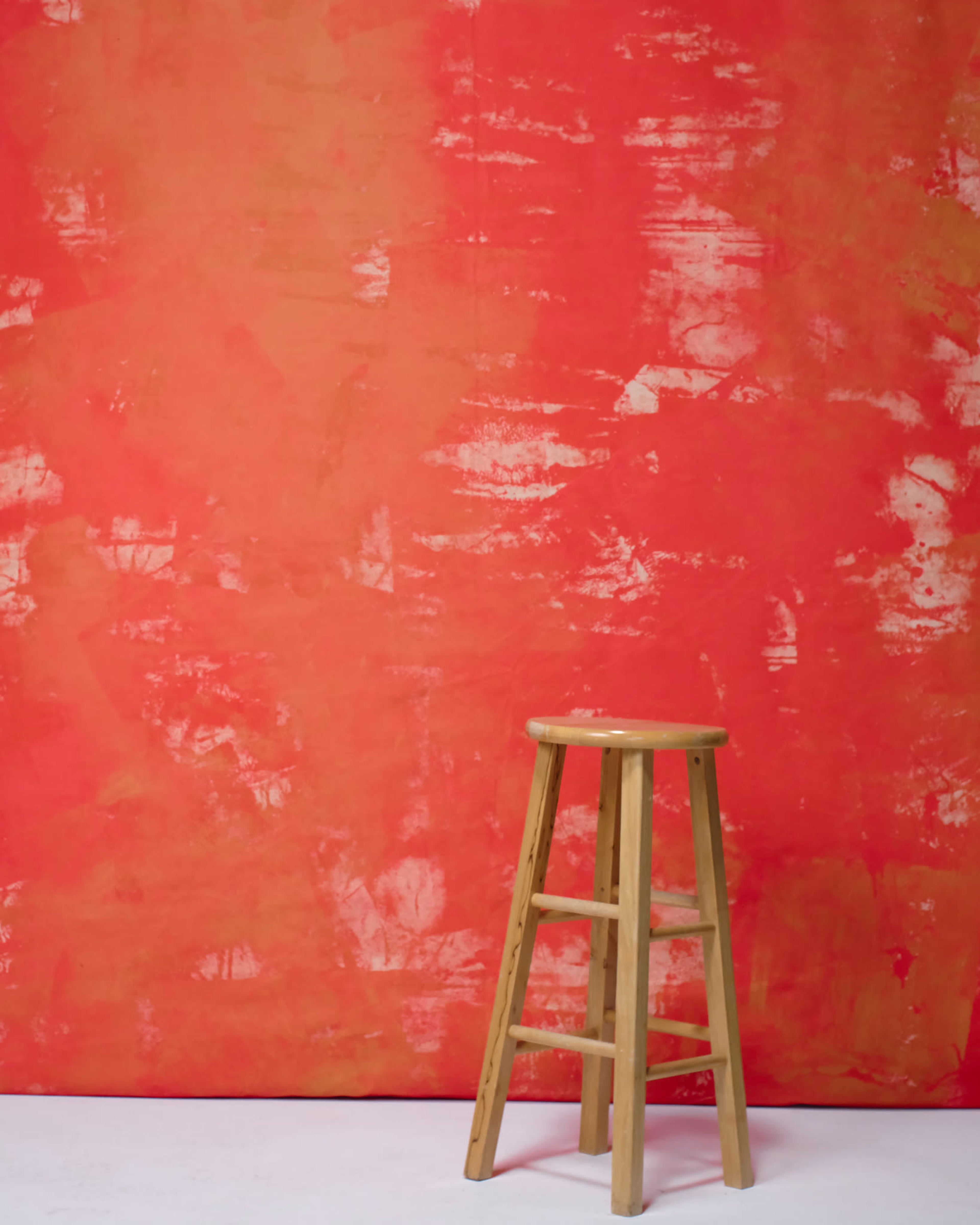 A wooden stool is positioned in front of a textured, bright red backdrop.