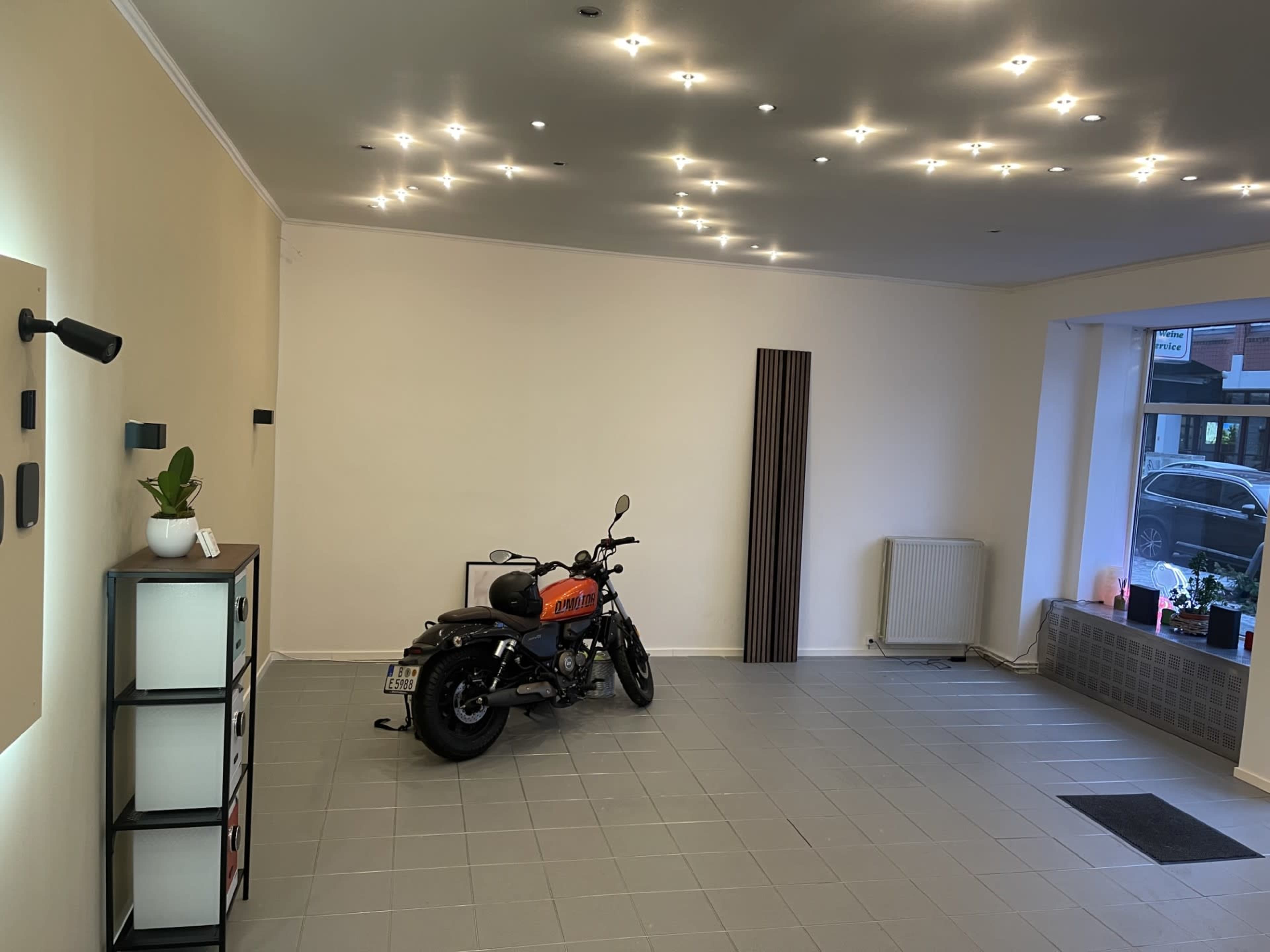 The image shows a spacious, well-lit room with a motorcycle parked in one corner and minimalist furniture along the walls.