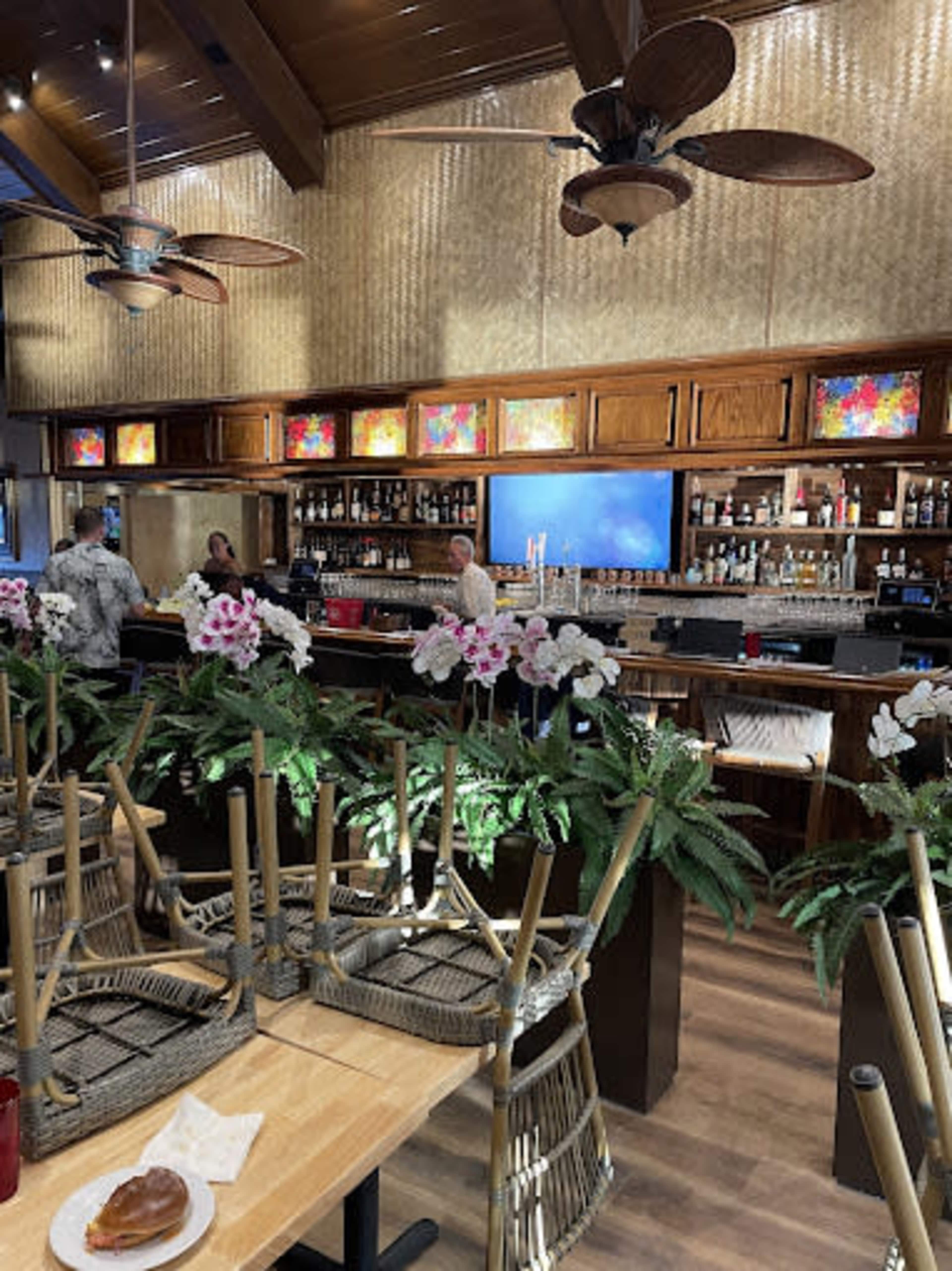 A bar area features wooden furnishings, tropical decor, and a counter lined with drink bottles, while tables are stacked with chairs.