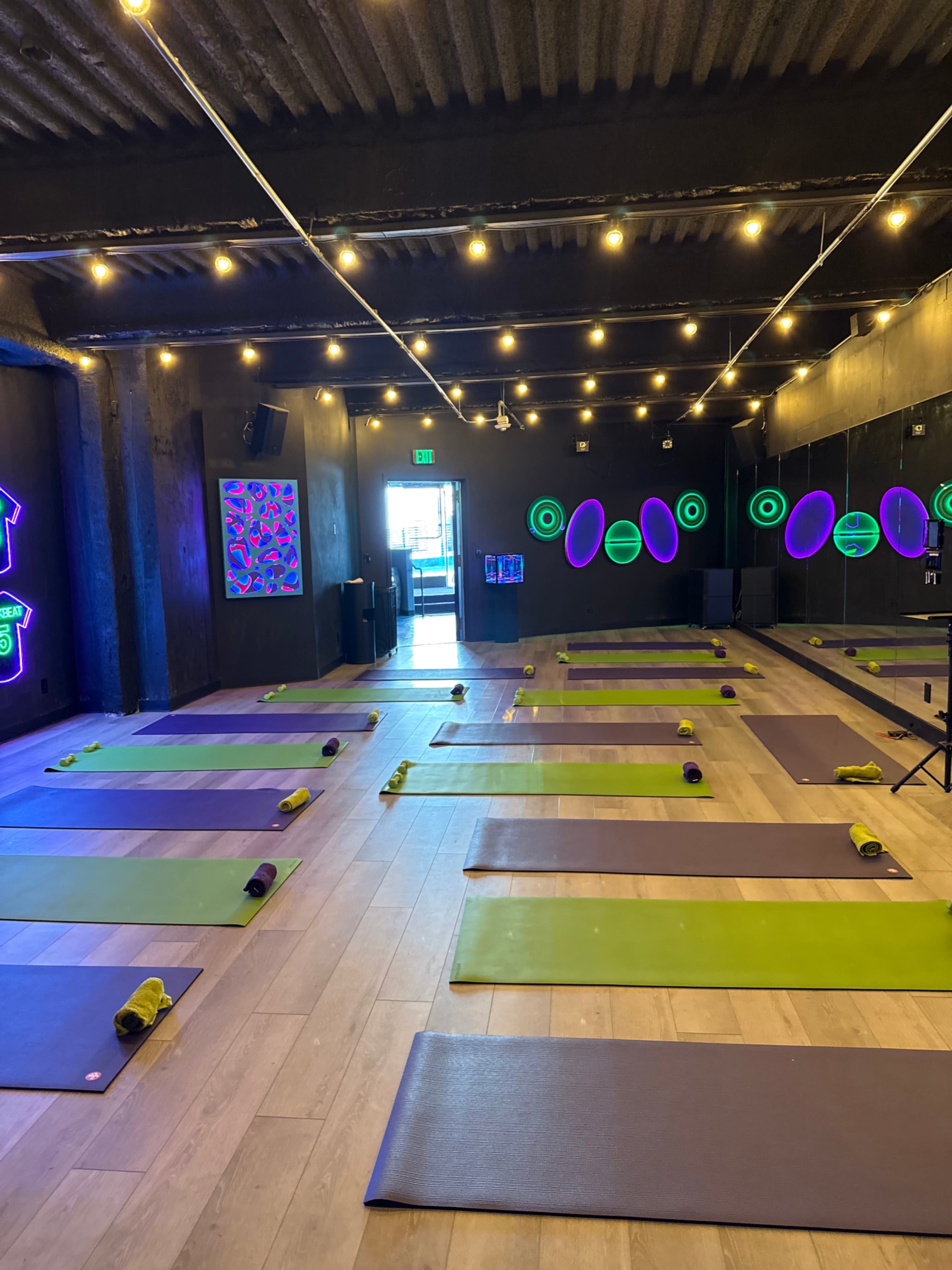 The image shows a yoga studio with multiple exercise mats arranged neatly on the floor, illuminated by colorful neon lights and string lights overhead.