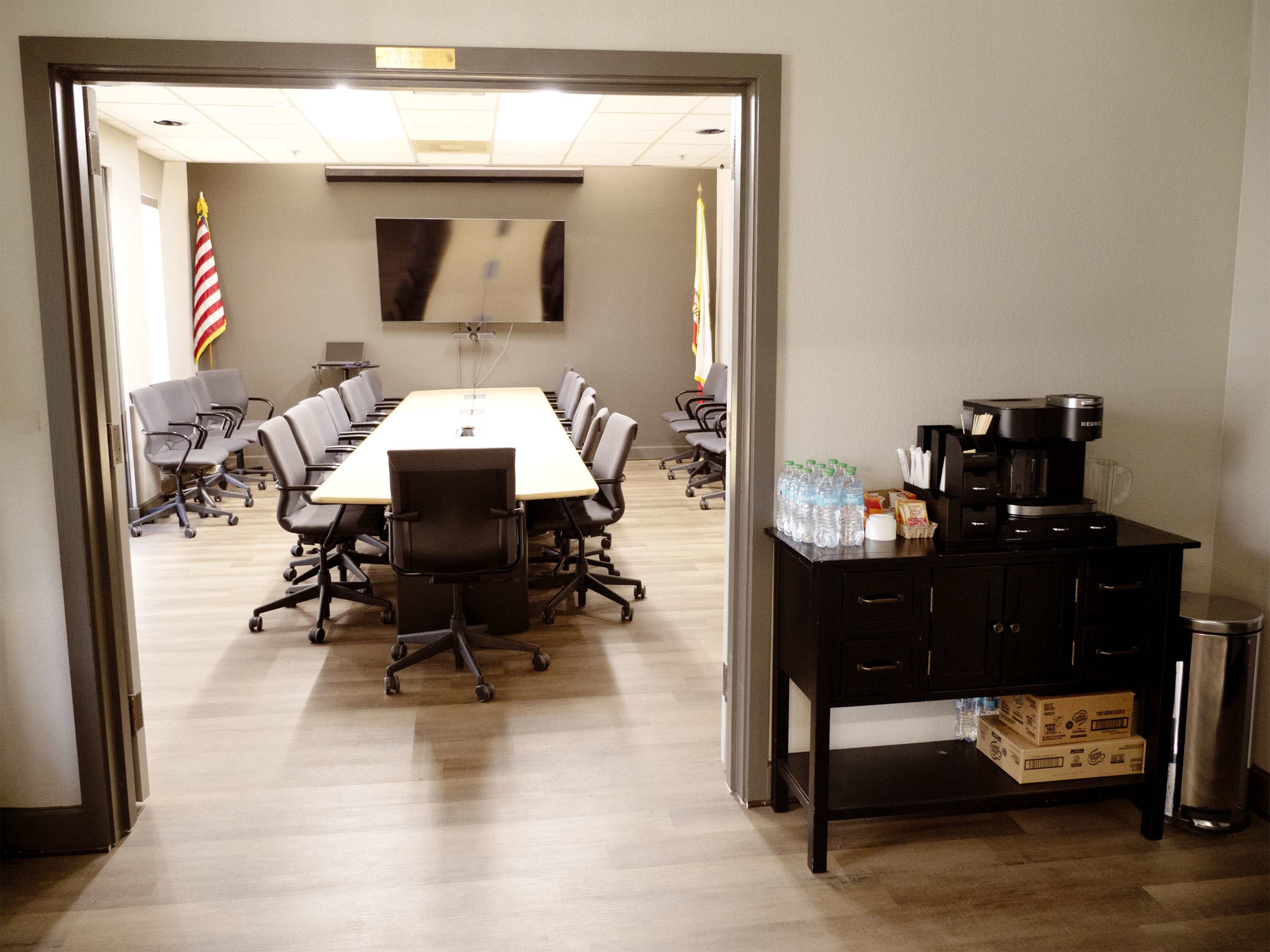 A conference room is seen through an open doorway, featuring a long table surrounded by chairs and a kitchenette area with a coffee machine and water bottles.