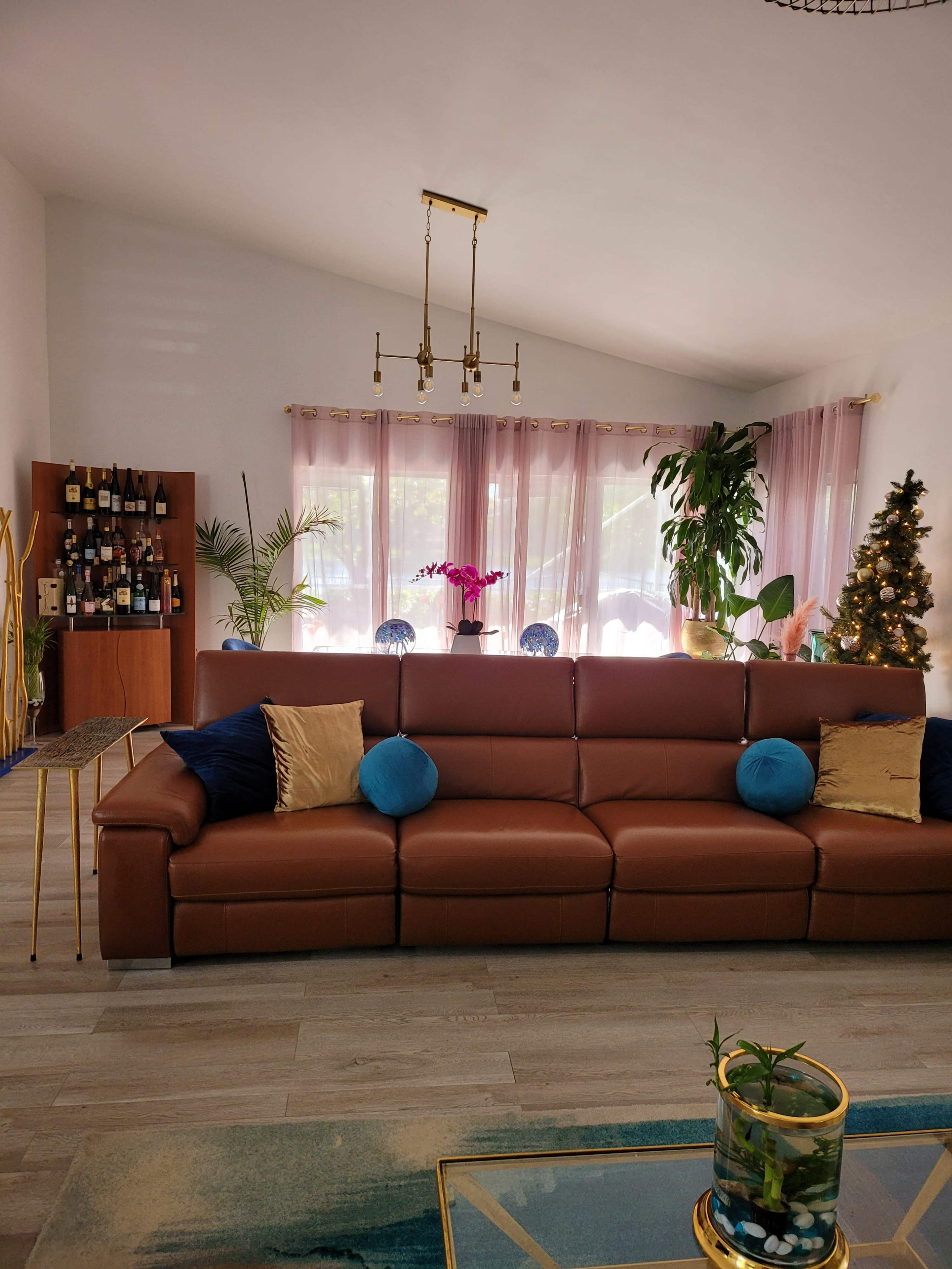 A modern living room featuring a brown sectional sofa with blue and gold cushions, a bar cabinet with a selection of drinks, and large windows dressed in sheer curtains.
