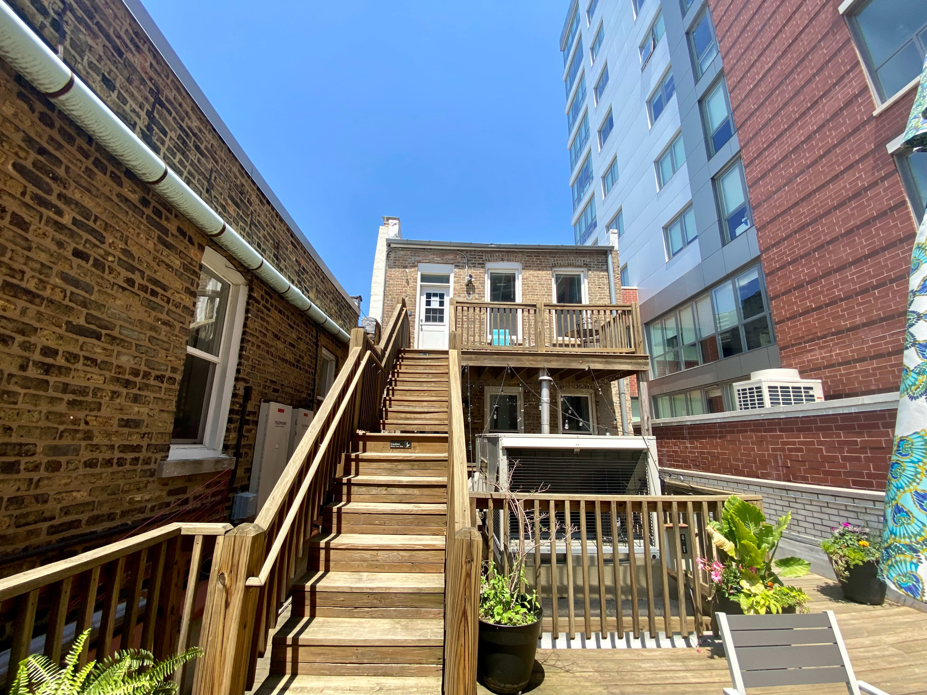 A wooden staircase leads to a second-floor deck of a brick building, surrounded by modern and older structures under a clear blue sky.