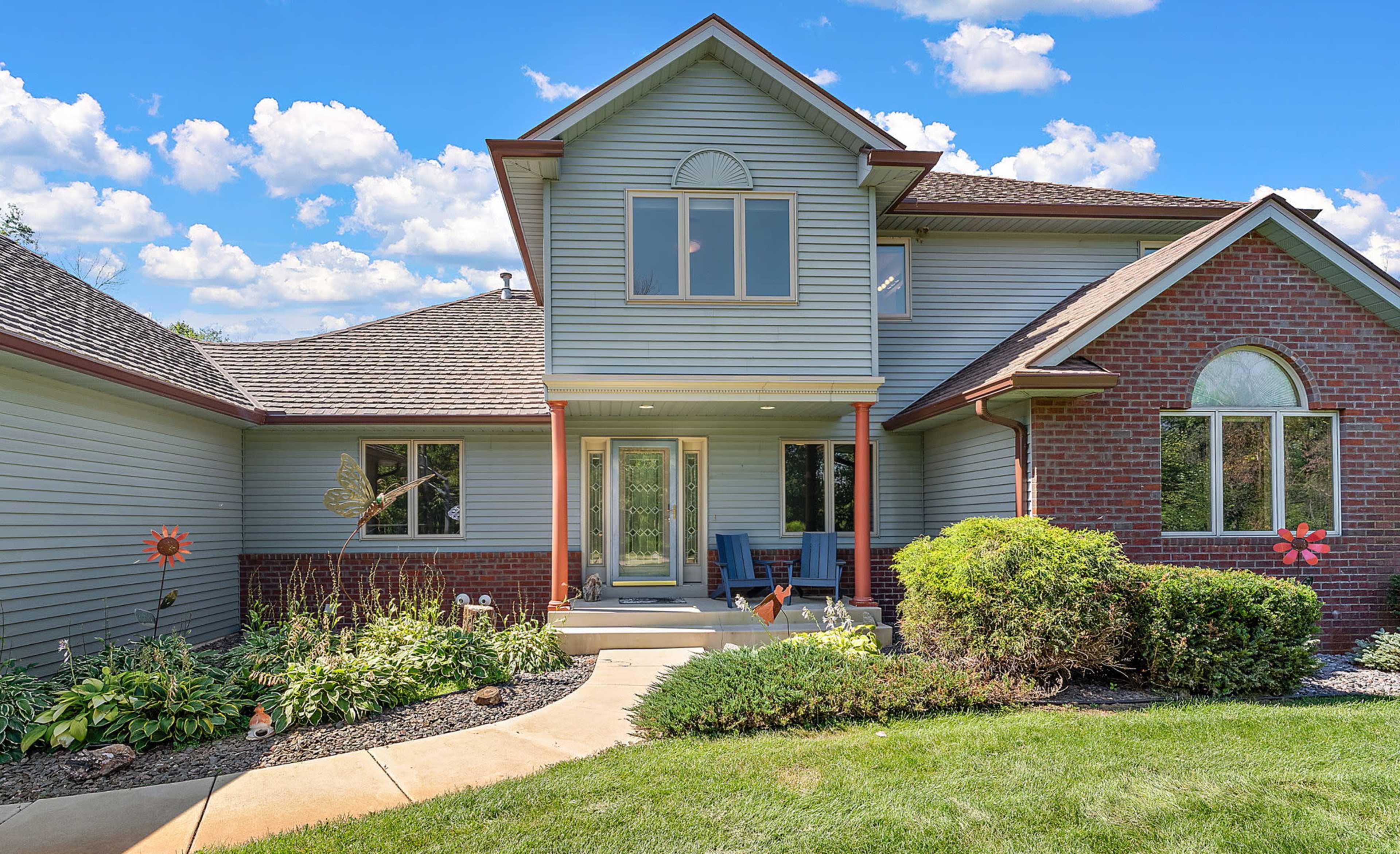 The image shows a two-story house with a combination of gray siding and brick, featuring a well-maintained front garden and a pathway leading to the entrance.