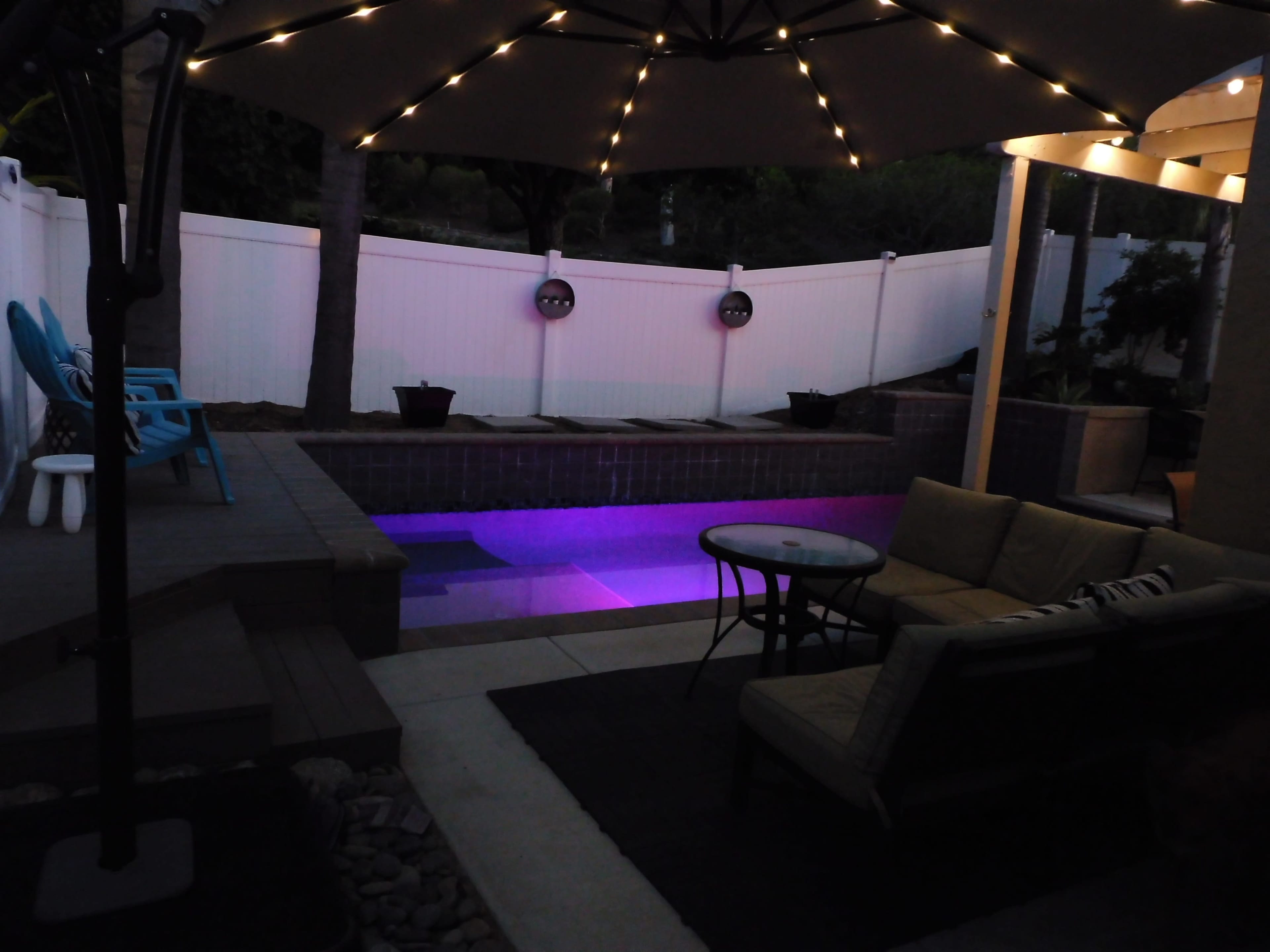 A backyard space features a pool with colored lights, a seating area with a table and chairs, and an umbrella overhead.