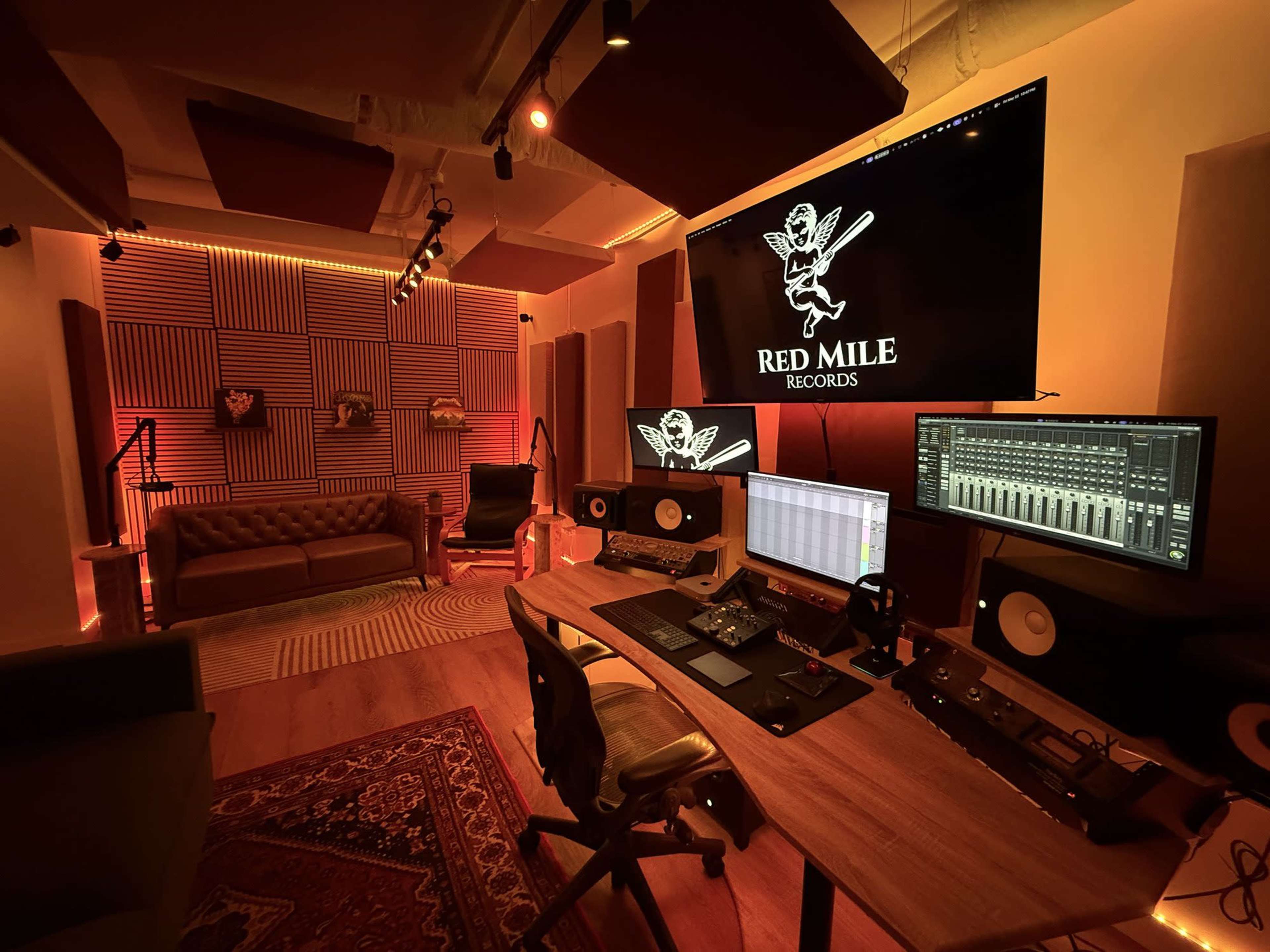 The image shows an interior of a recording studio featuring a cozy seating area, a large monitor displaying the "Red Mile Records" logo, and audio equipment on a desk.