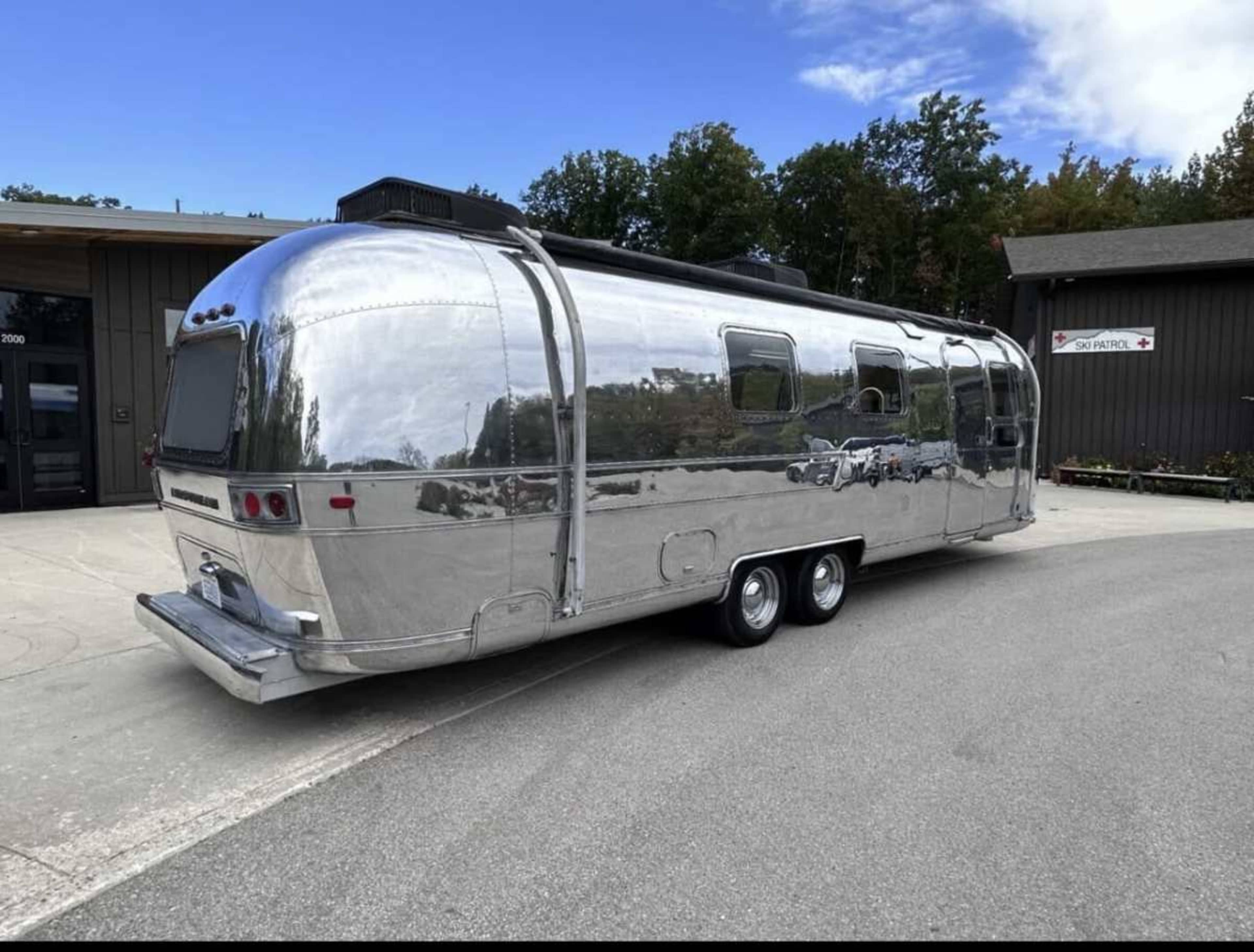A shiny silver Airstream travel trailer is parked on a paved area next to a building with green trees in the background.