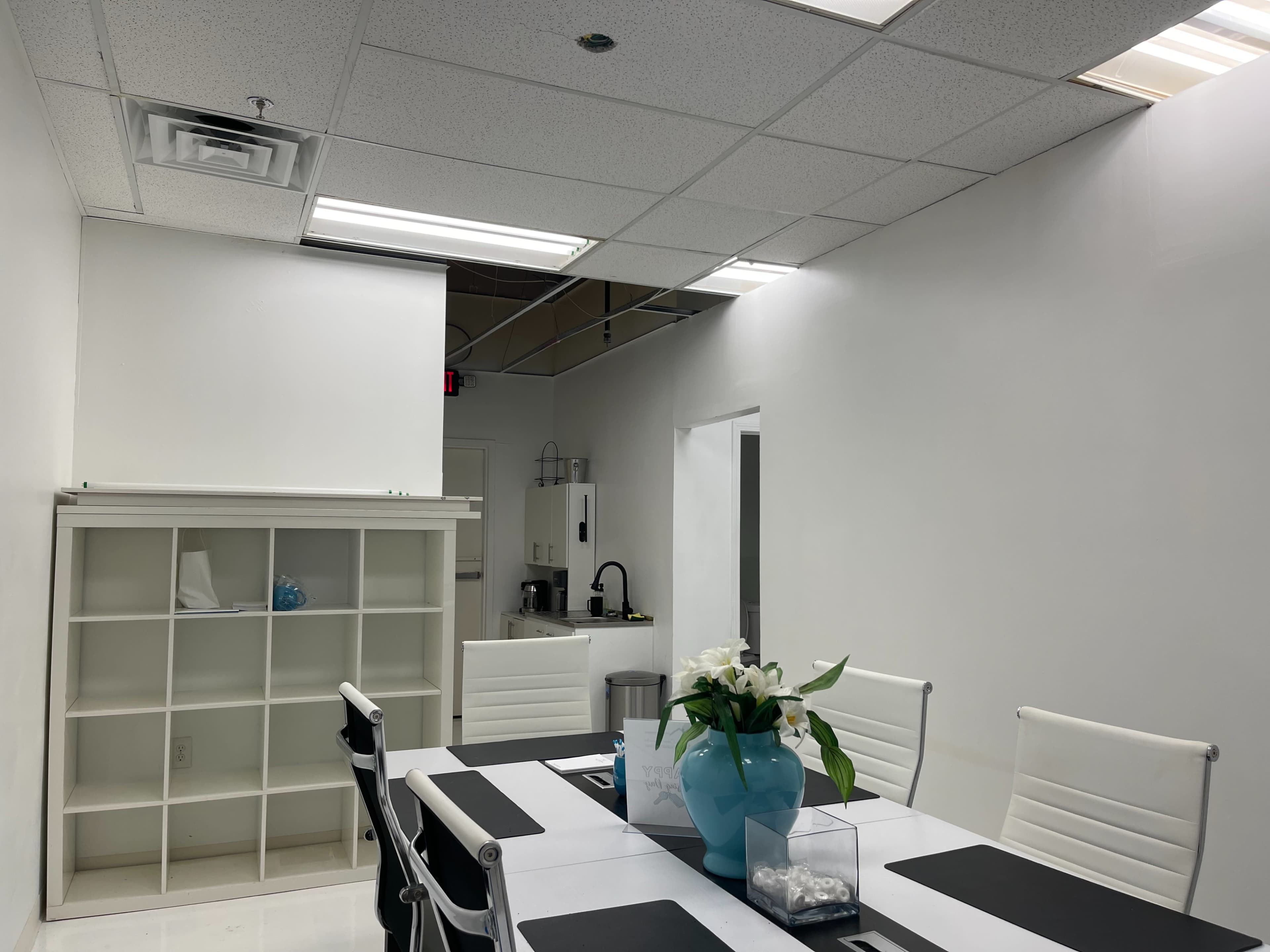 A modern meeting room features a white table surrounded by chairs, with a blue vase of flowers in the center and a kitchenette area in the background.