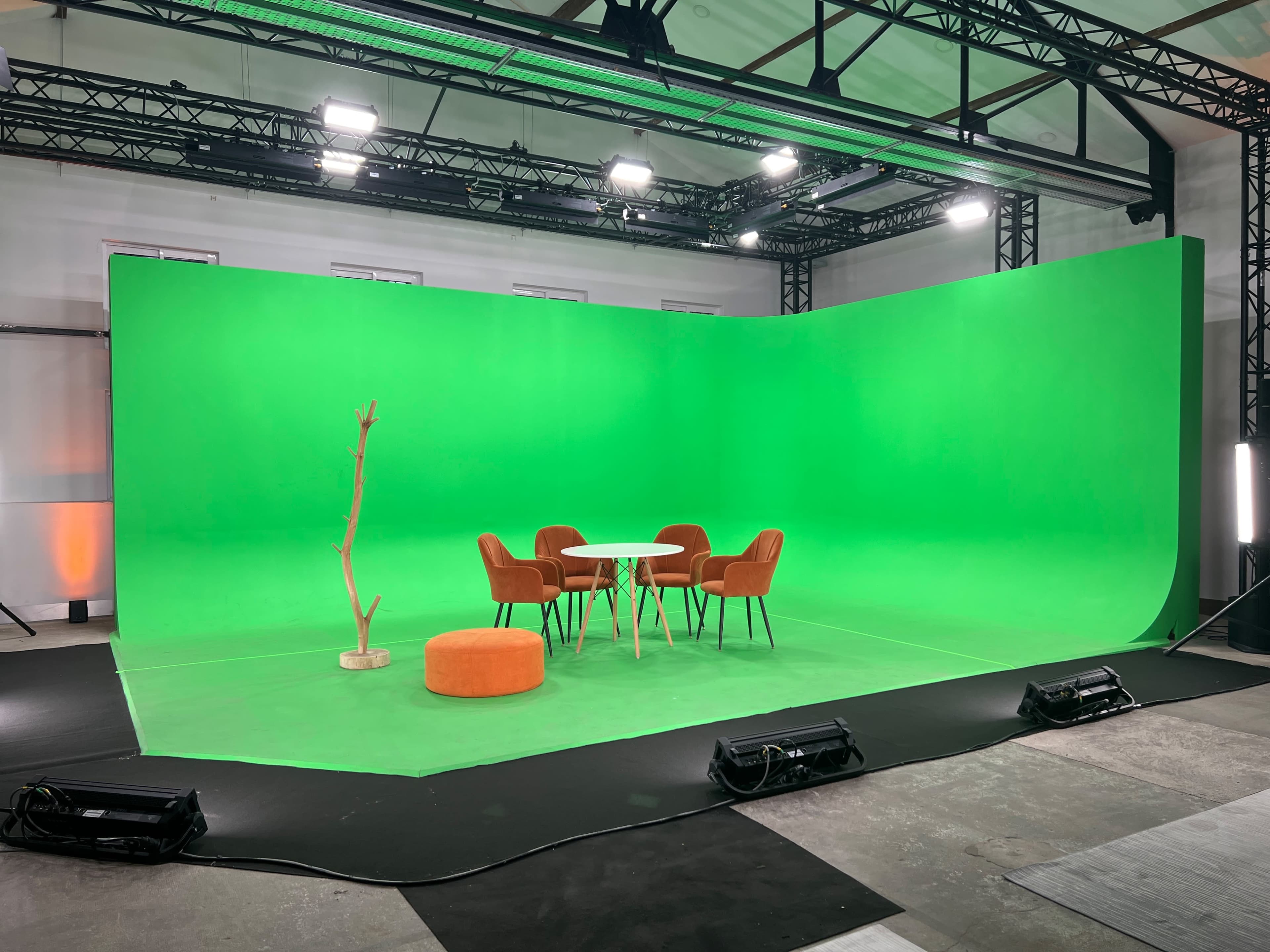 A green screen studio featuring a circular orange seating, a small white table, and four brown chairs arranged under overhead lighting.