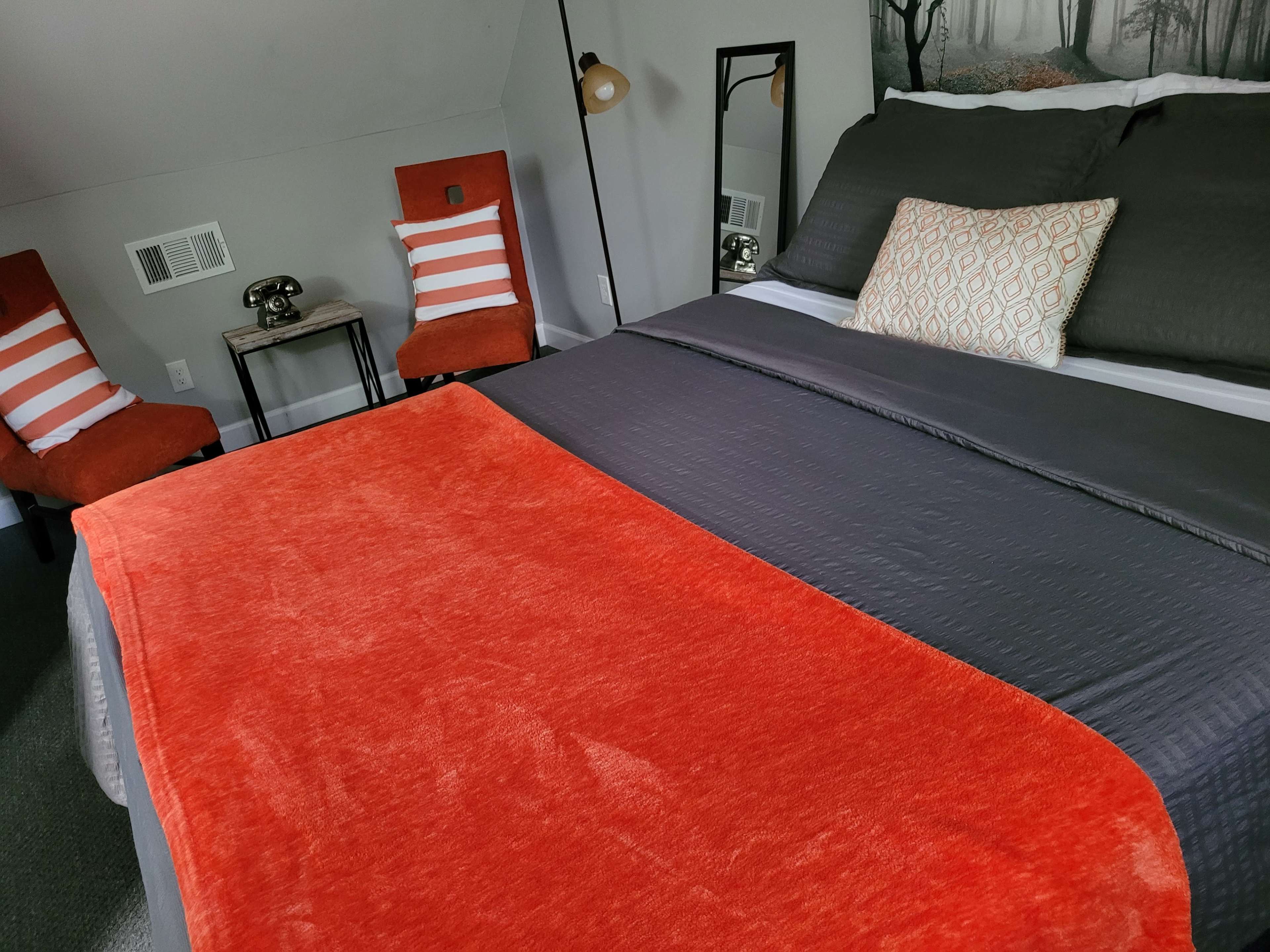 A neatly made bed with an orange blanket, flanked by two red chairs and a mirror in a bedroom with a forest-themed wall decor.