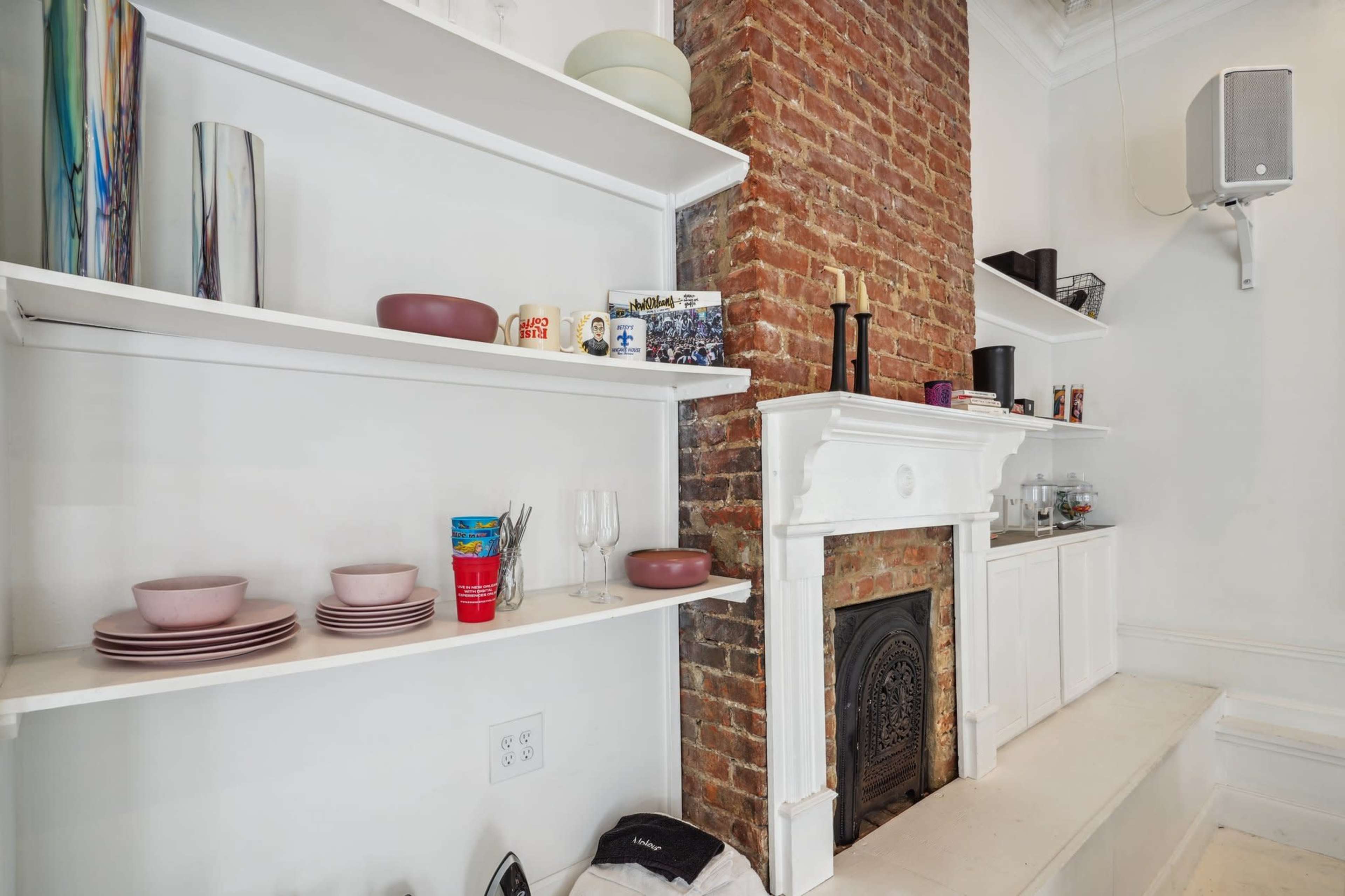 A white shelf holding dishware and glassware is positioned next to a brick fireplace in a well-lit room.