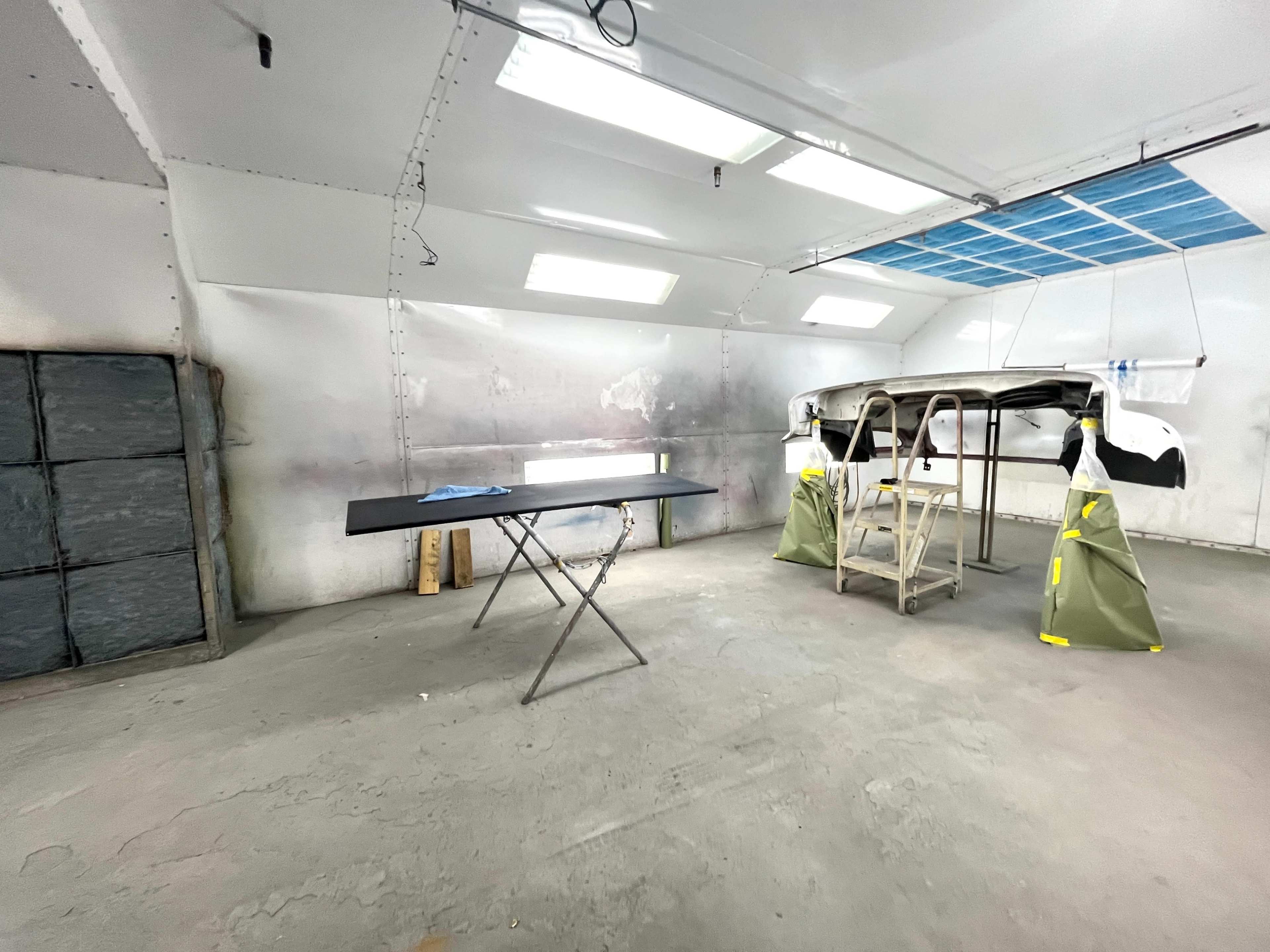 The image shows an empty industrial workspace with a folding table in the center and a car part mounted on stands in the background.