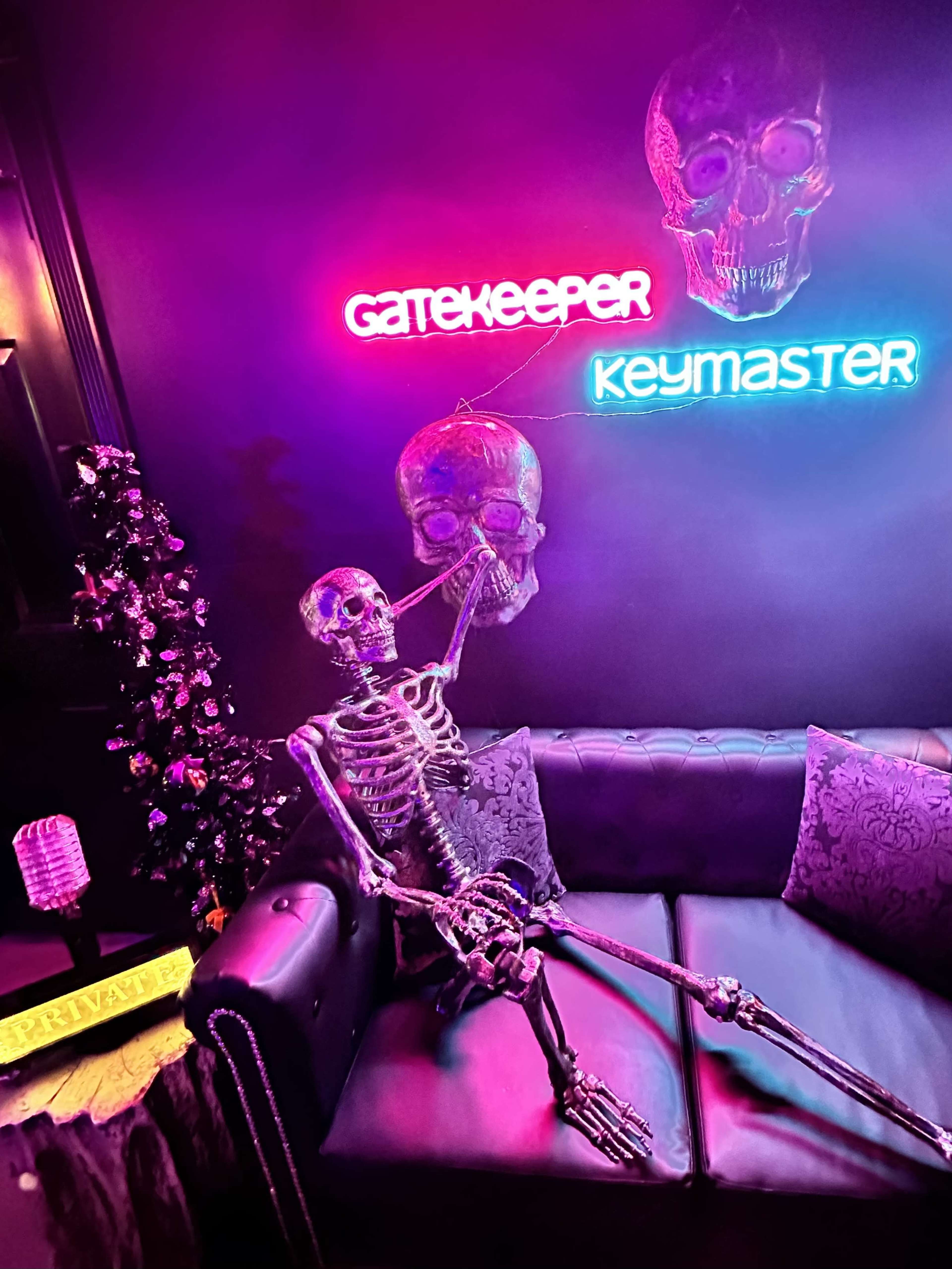 A skeleton is seated on a black couch in a dimly lit room adorned with neon signs that read "GATEKEEPER" and "KEYMASTER."