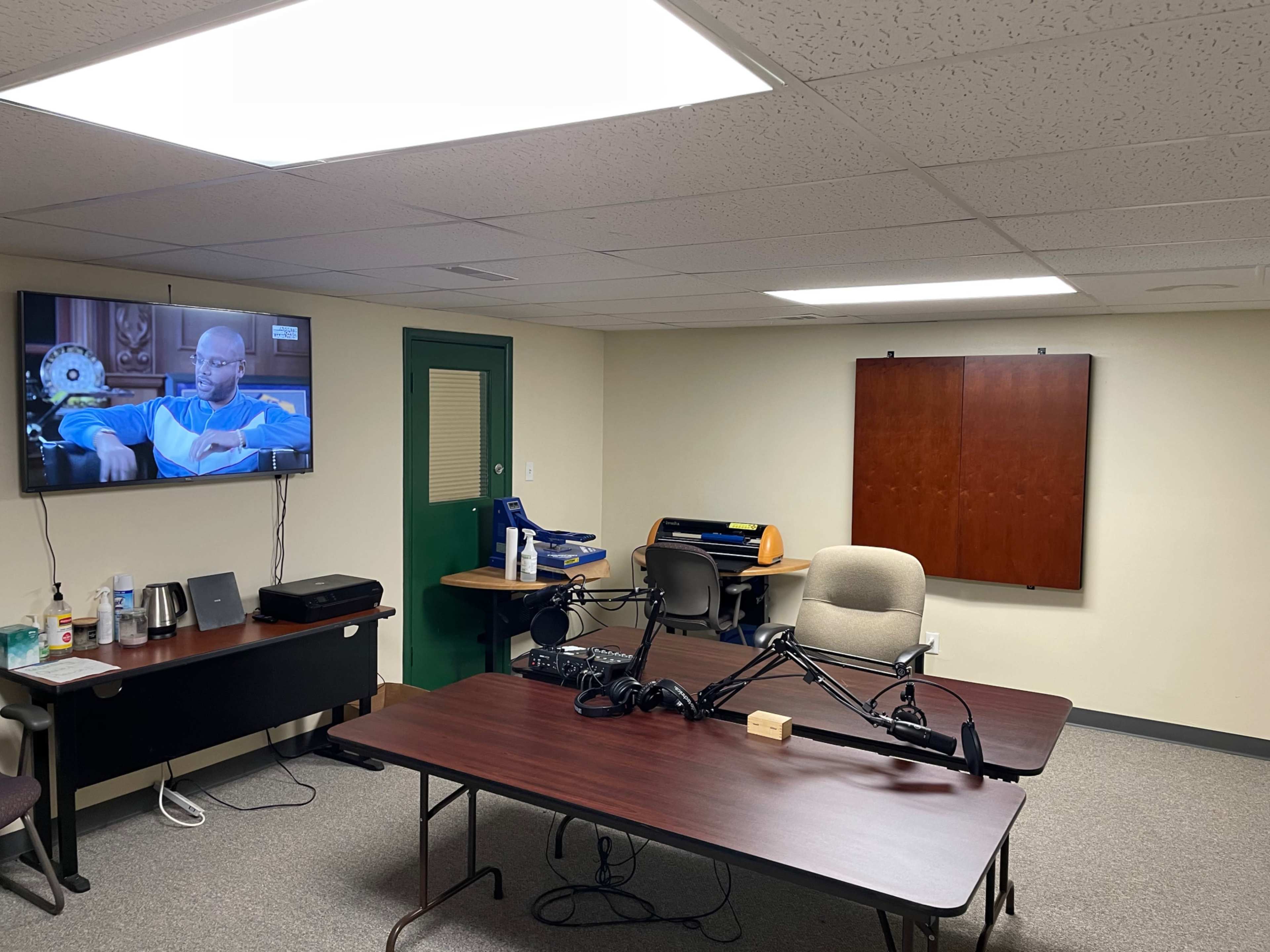 The image shows a sparsely furnished meeting room with a large television on the wall, a desk with office supplies, and a table equipped for recording or broadcasting.