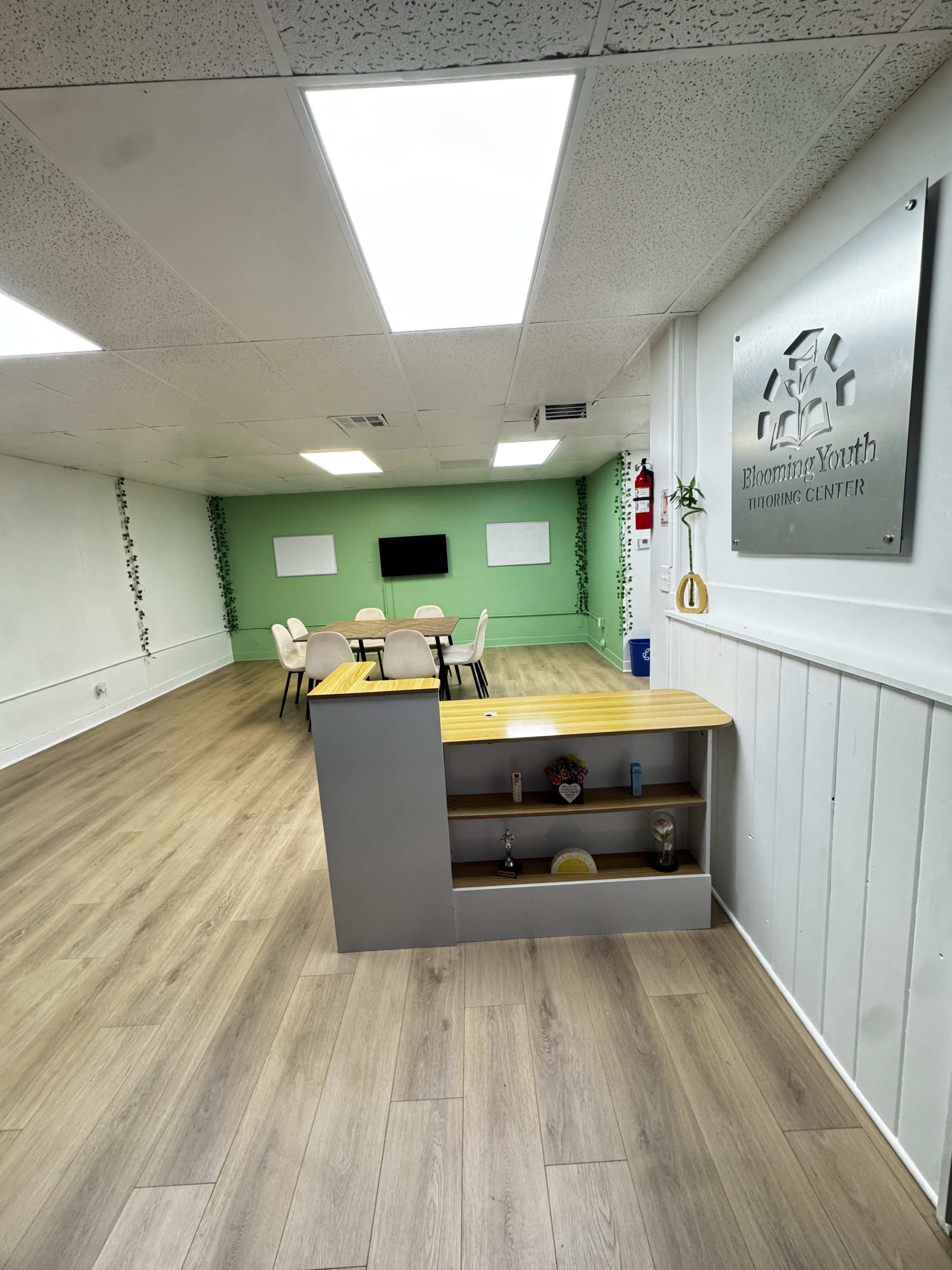 The image shows a spacious room with light wood flooring, a reception area on the left, and a meeting space with a green accent wall and a table surrounded by chairs in the background.