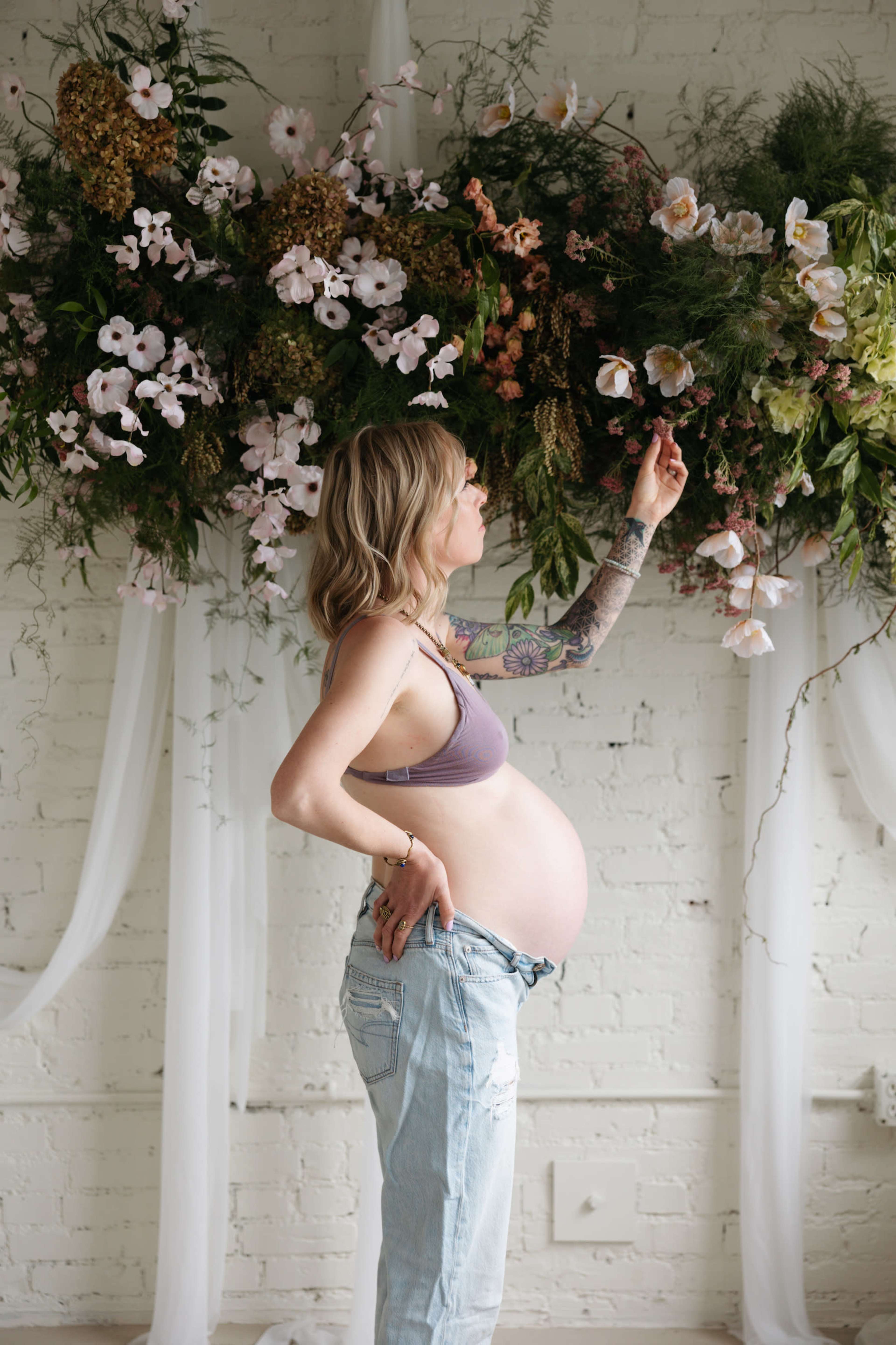 A pregnant woman stands in profile, touching a floral arrangement mounted on a wall while wearing casual clothing.