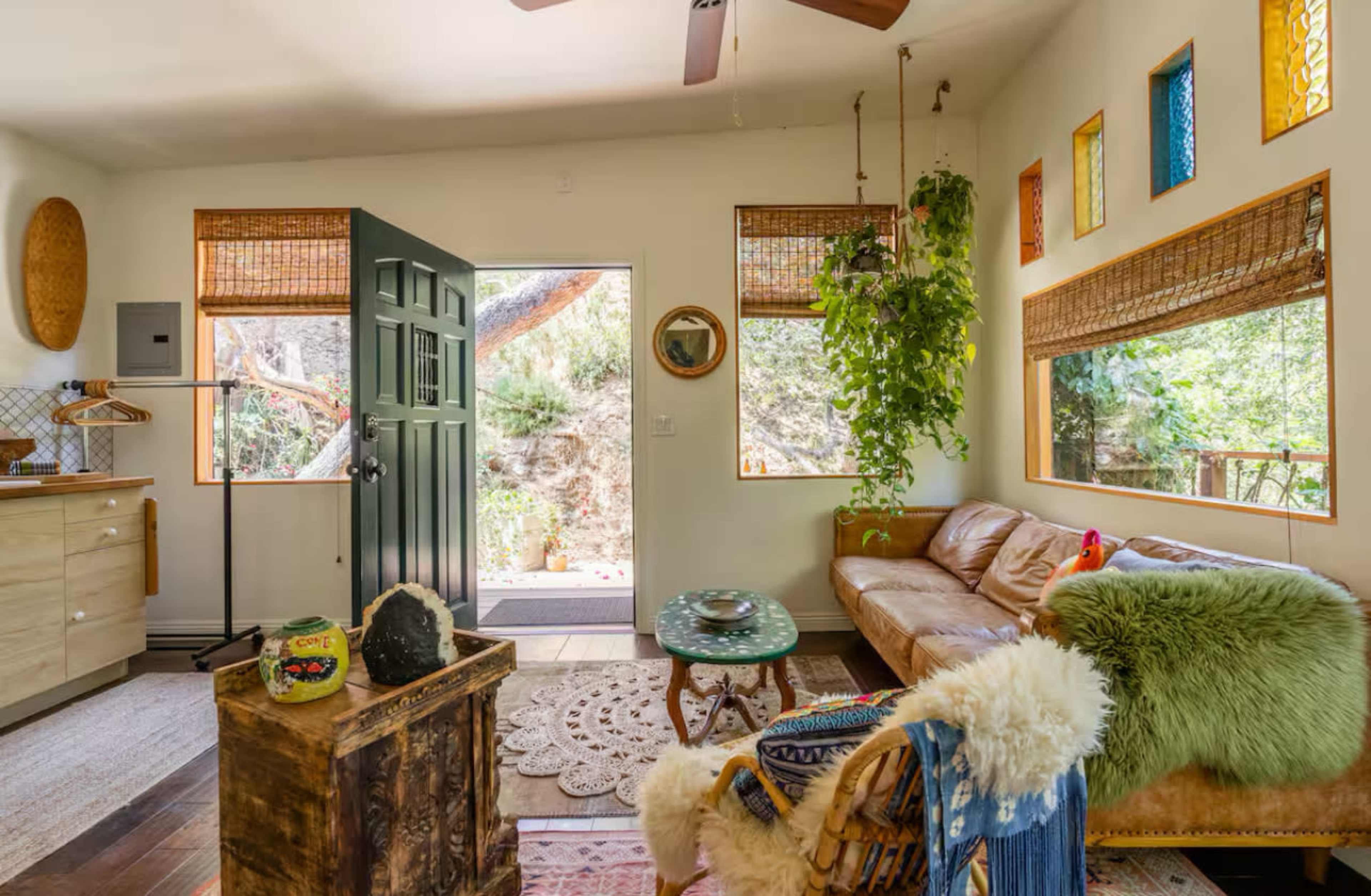 The image shows a cozy living room with a brown leather couch, potted plants, and large windows that open to a natural outdoor setting.