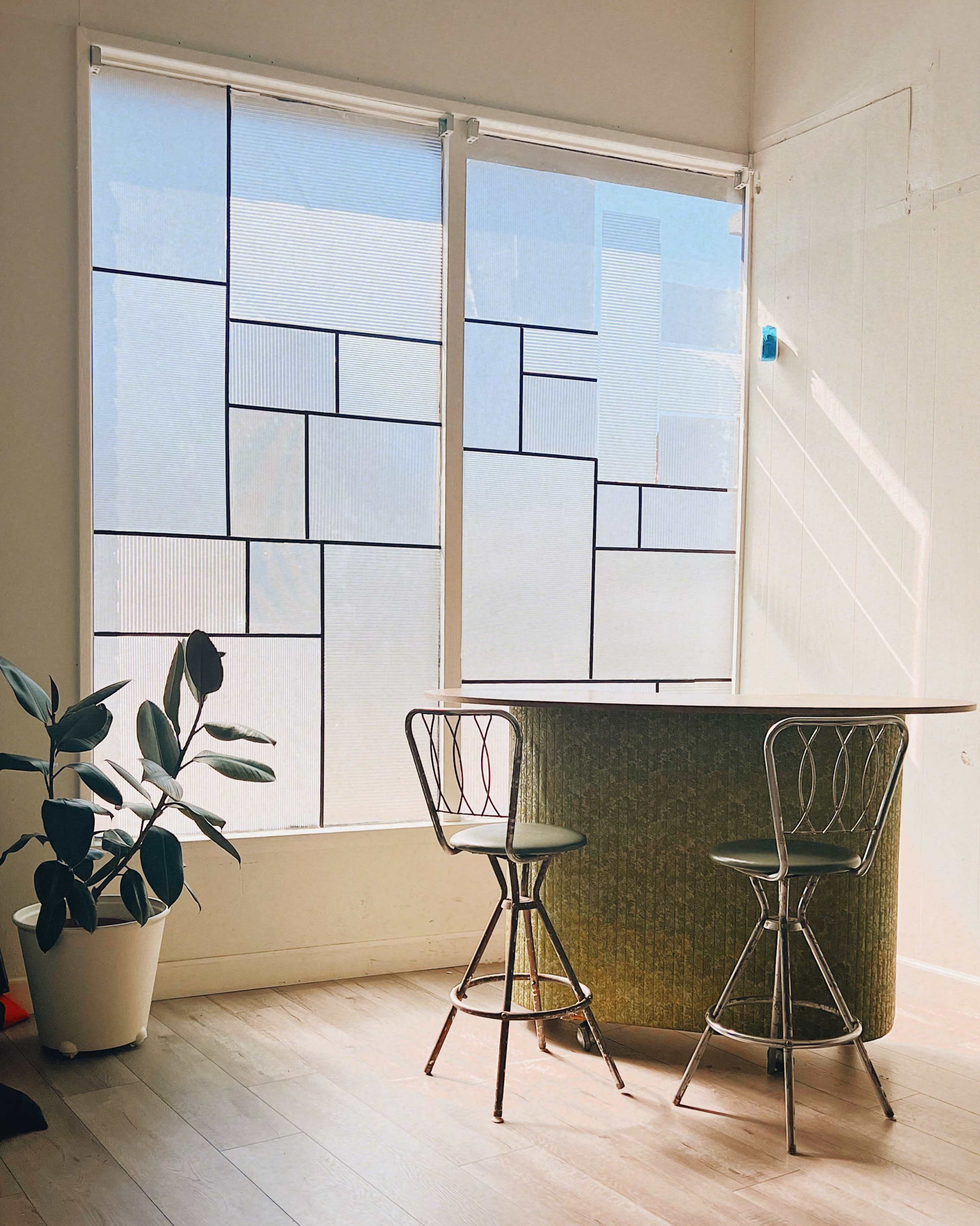 The image features a modern interior with a green bar counter, two metal stools, and large windows covered with patterned sheer blinds.