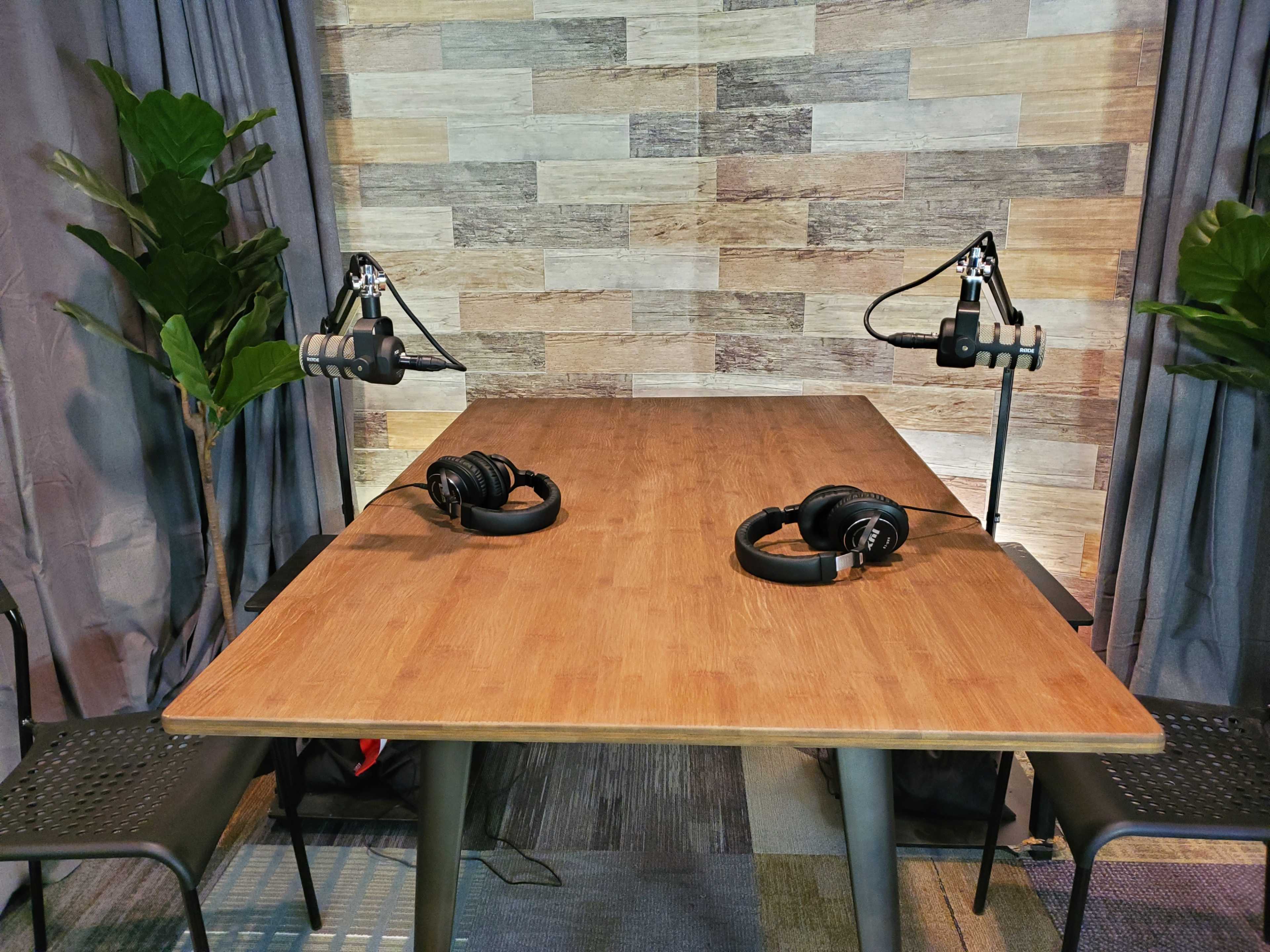 A wooden table with two microphones and headphones is set up in a room with a wooden panel background and potted plants on either side.