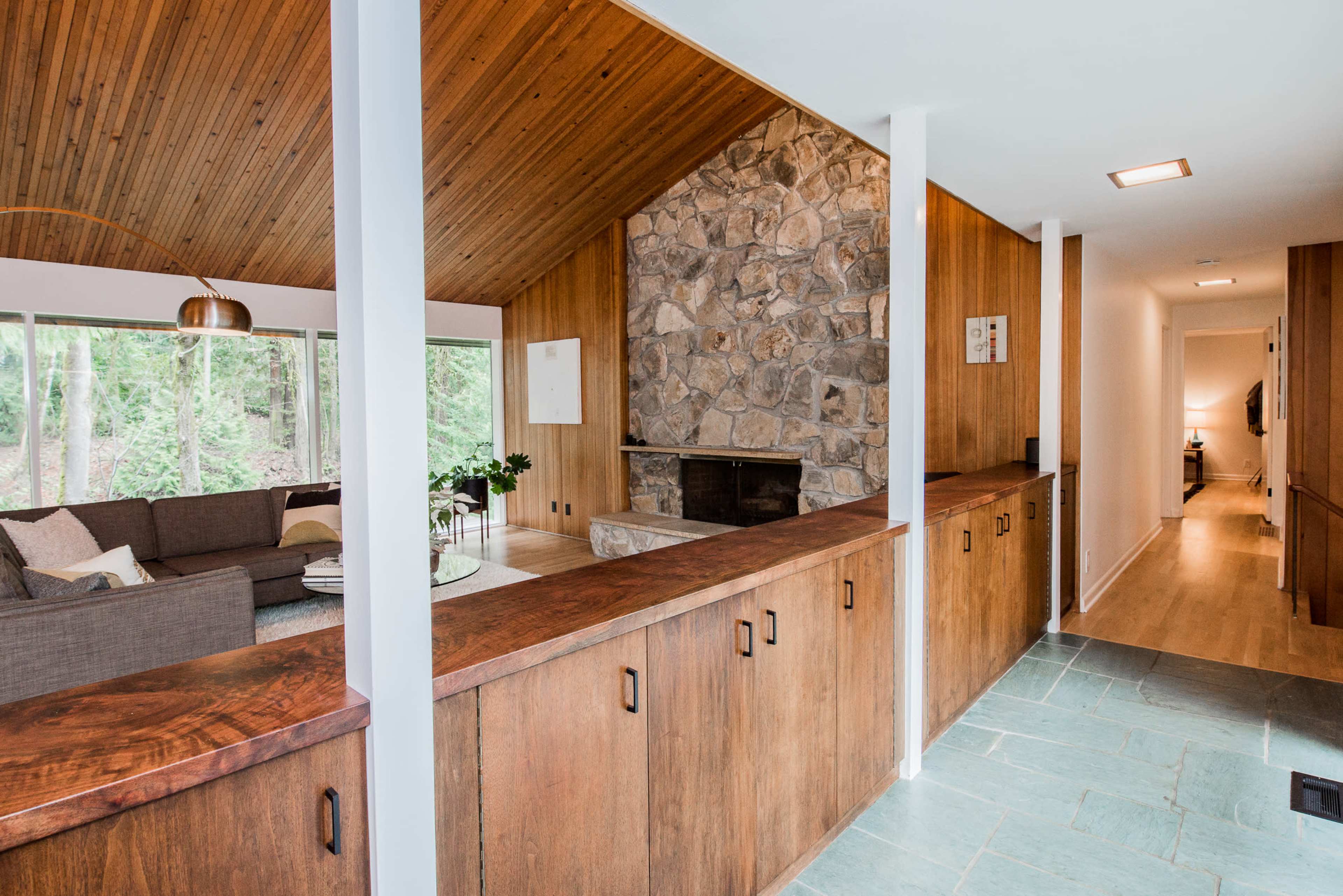 A modern living room with a stone fireplace, wooden paneling, and large windows that overlook a forested area.