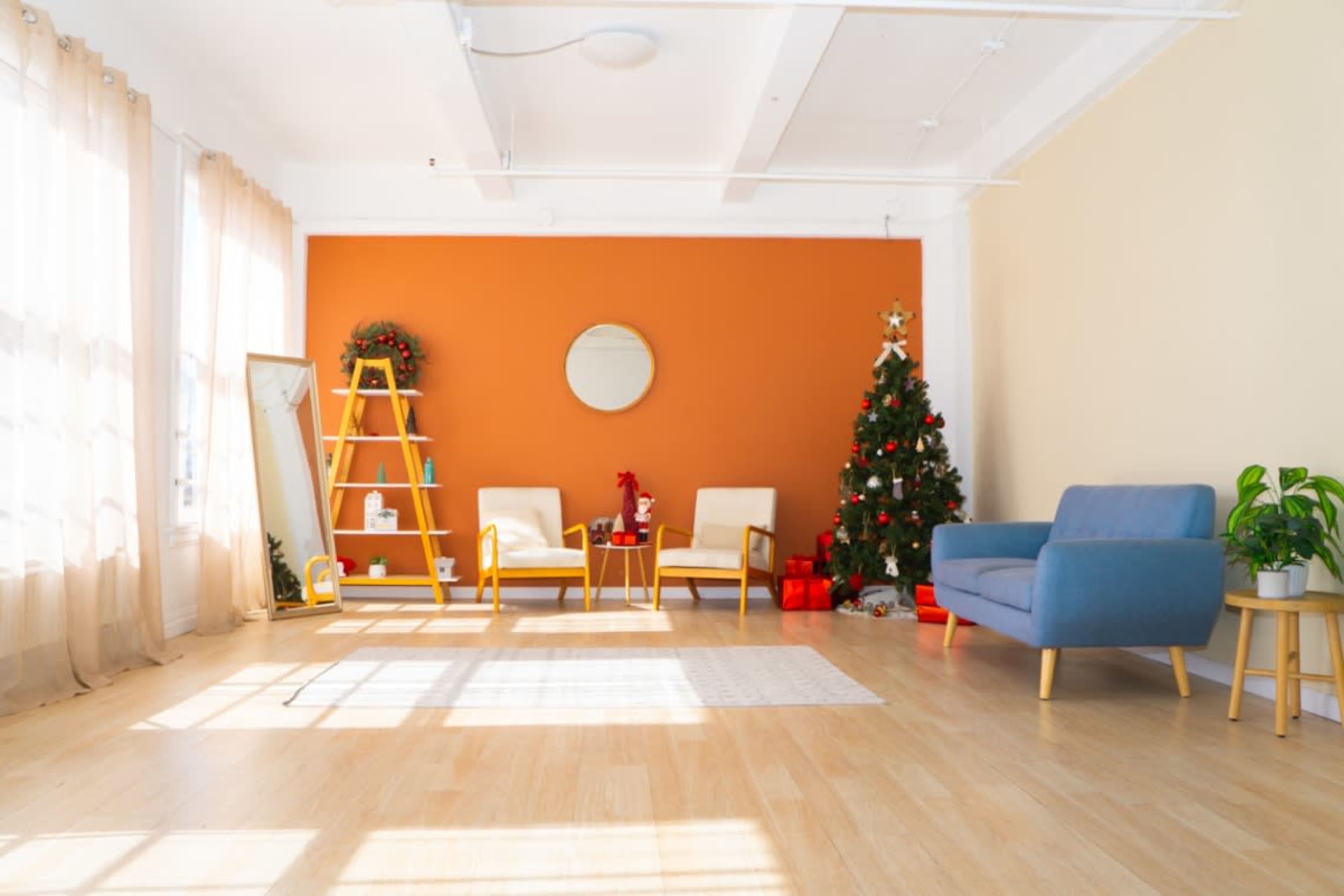 The room features a decorated Christmas tree against an orange wall, with seating and a mirror arranged around it, and large windows allowing natural light to fill the space.