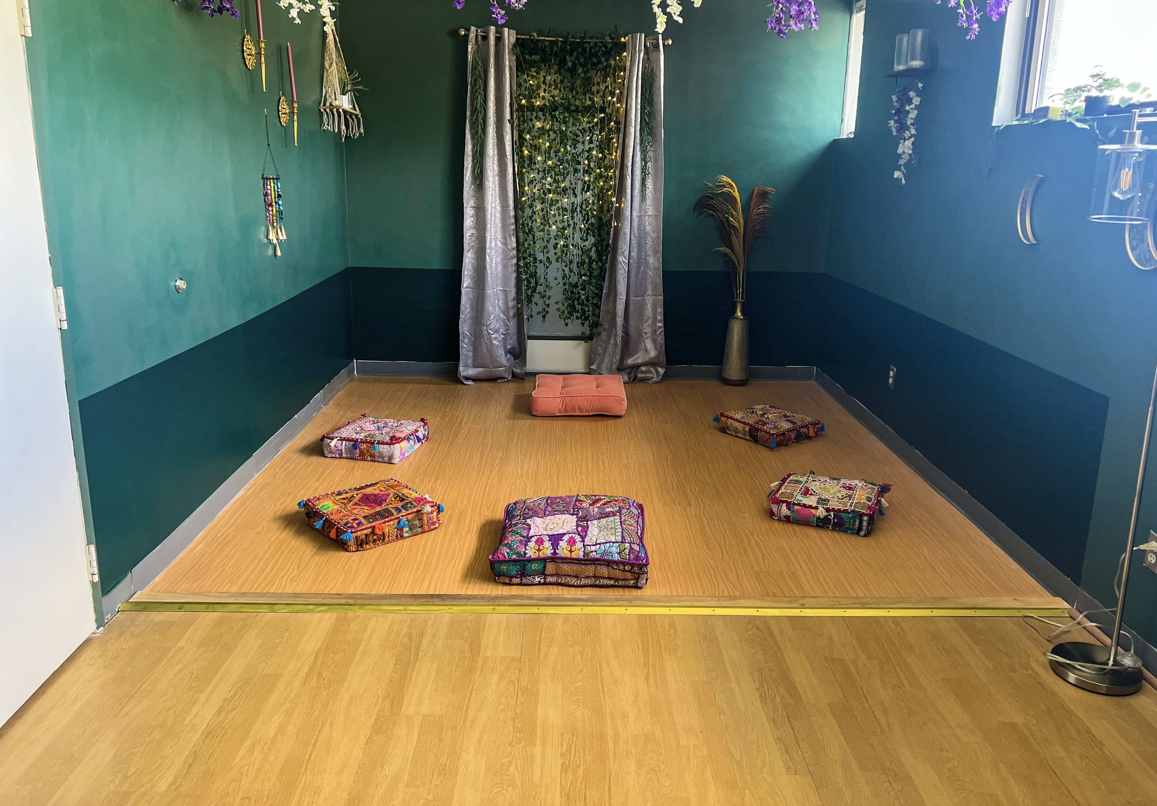 The image shows a spacious room with green walls, decorated with plants and cushions arranged on a wooden floor.