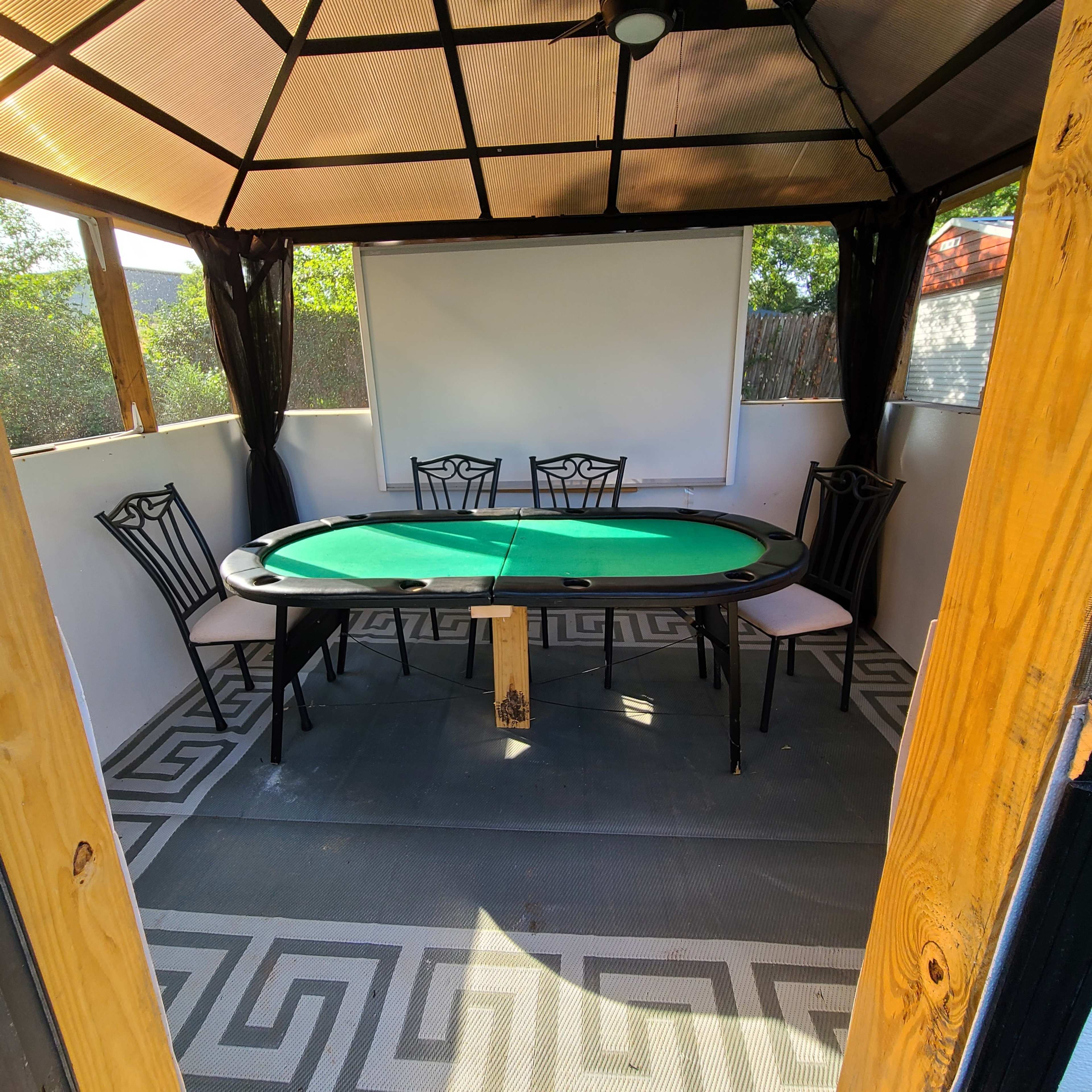 A gazebo-style structure containing a green felt poker table surrounded by four black chairs and a blank screen.