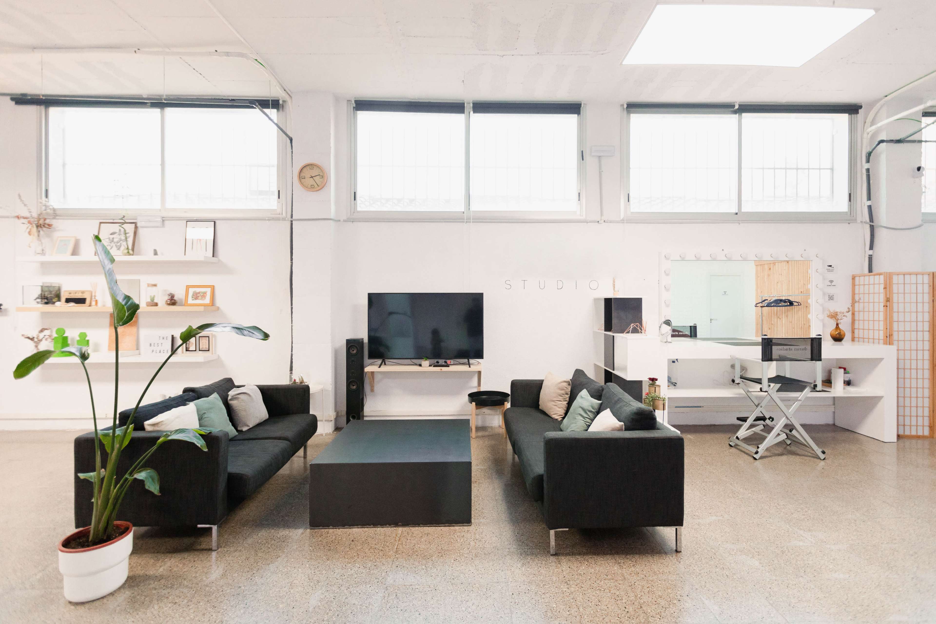 An open studio space with two black sofas facing a coffee table, a large television, and a mirror setup in a well-lit room with large windows.