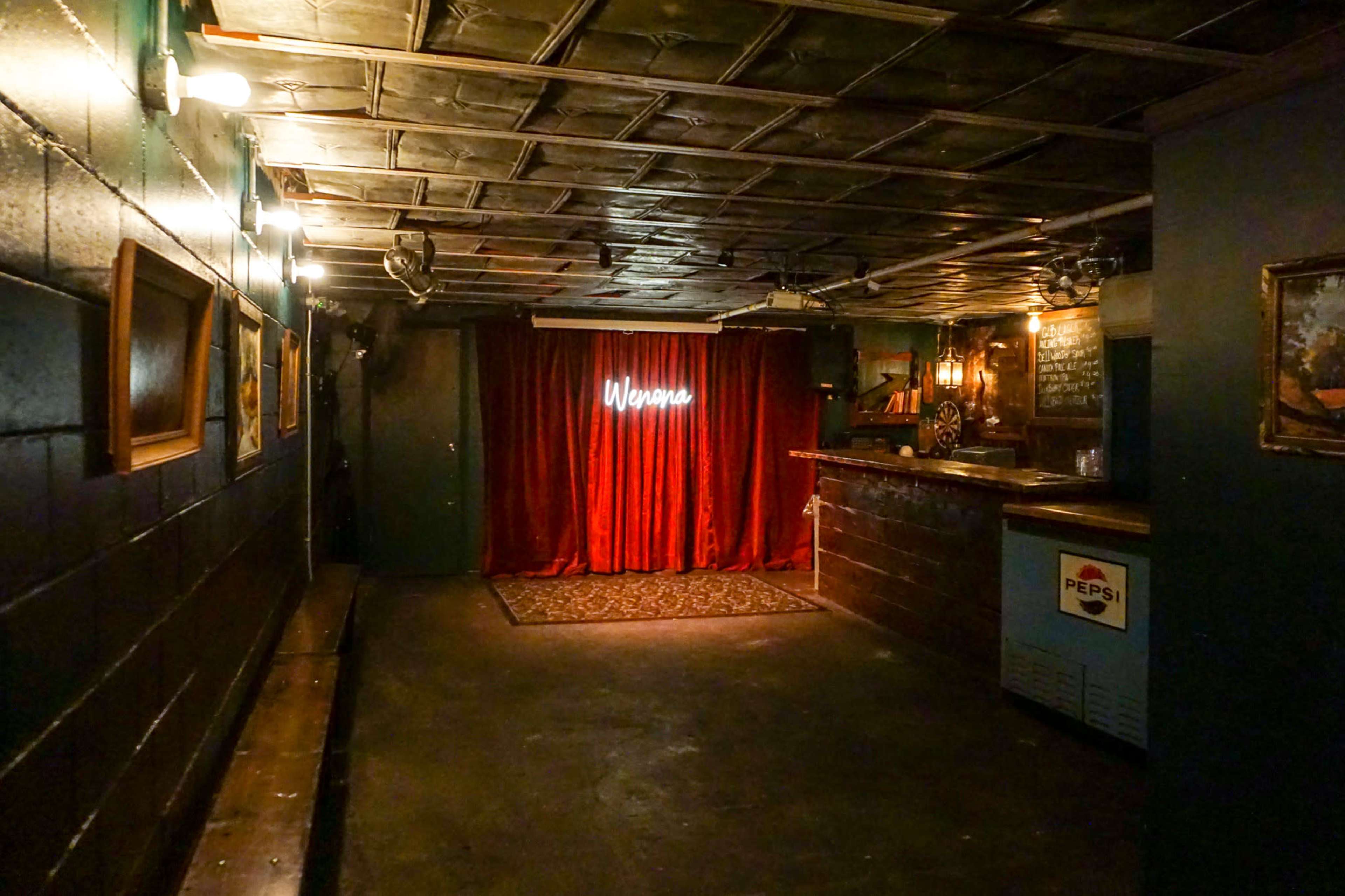An empty room features a red velvet curtain in front of a small stage, with a bar and dim lighting along the walls.