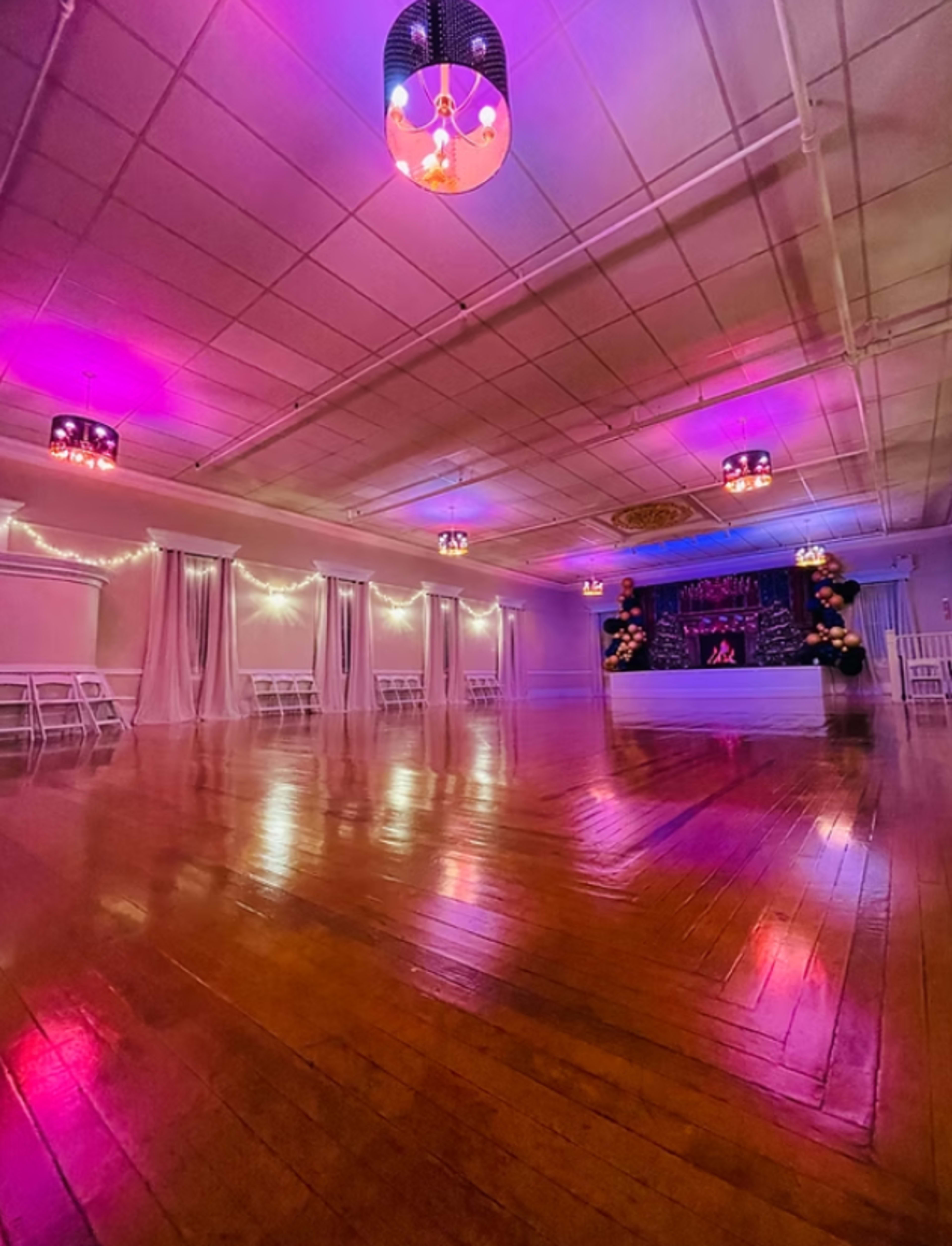 The image shows an event space with polished wooden floors, decorated with purple and pink lighting, white chairs, and a stage area at one end.