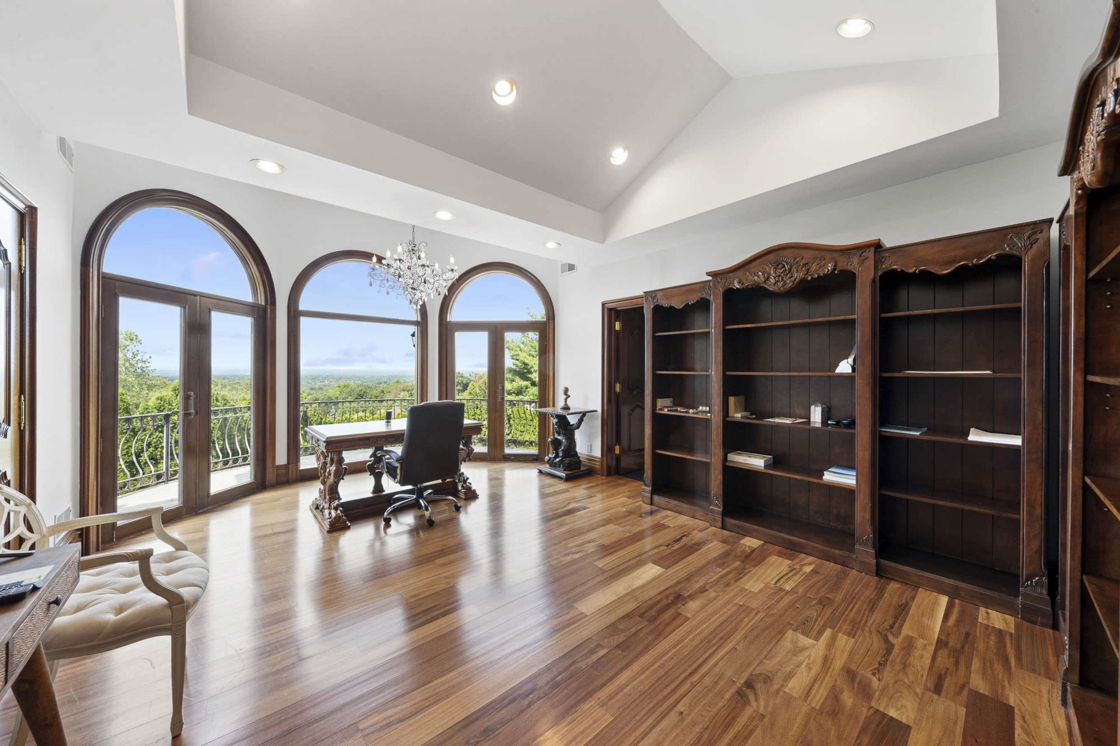 A wood-floored office space with large arched windows, a chandelier, a desk, and a bookshelf.